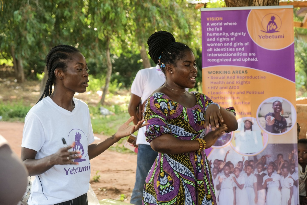 Two women speaking beside a Yebetumi banner in Ghana