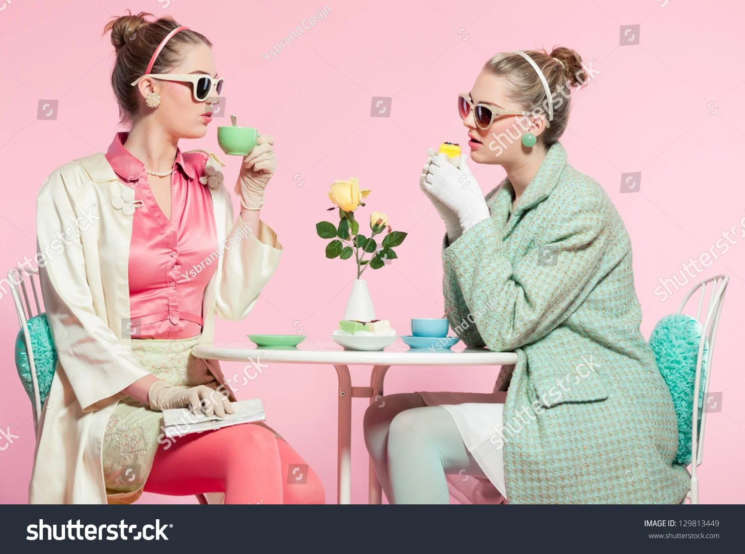 Two women dressed in vintage 1950s style sitting at a table with a pink background, drinking tea and engaging in conversation. One woman wears a pink satin blouse, cream coat, pink skirt, gloves, and sunglasses; the other in a mint-green patterned co