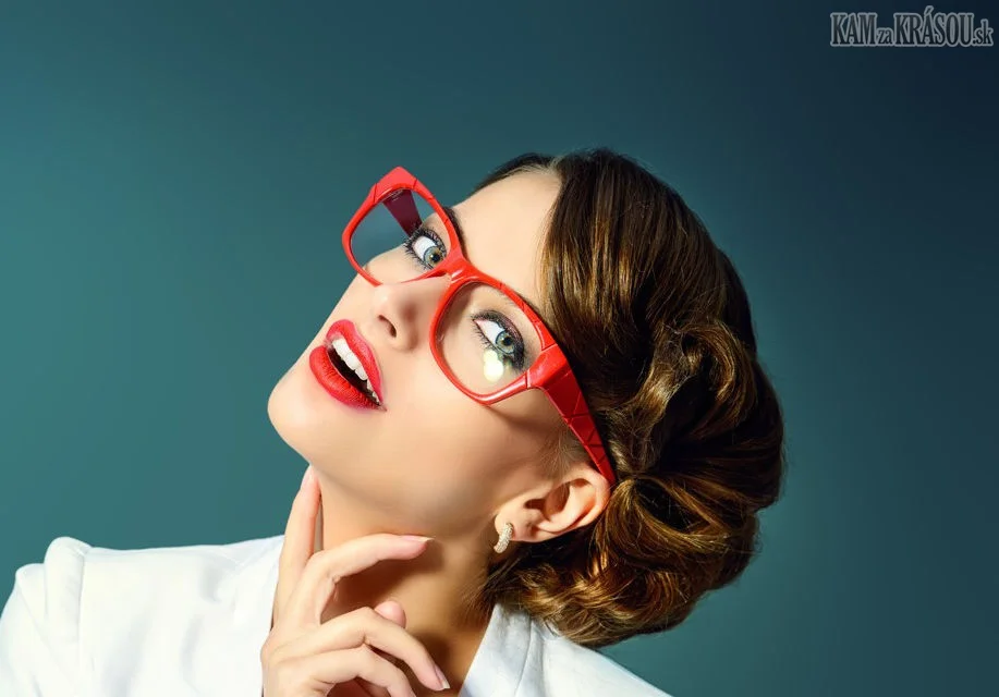 A woman with styled brown hair and red glasses posing with her head tilted back, touching her chin with her hand, wearing a white top, and looking upwards.