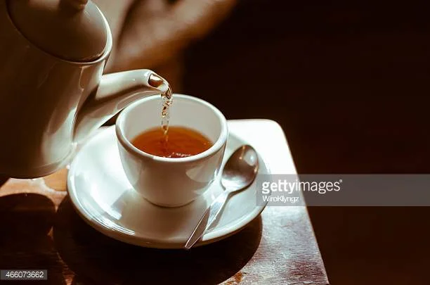 A white teapot pouring tea into a white teacup on a saucer with a silver spoon resting on it, placed on a wooden table.