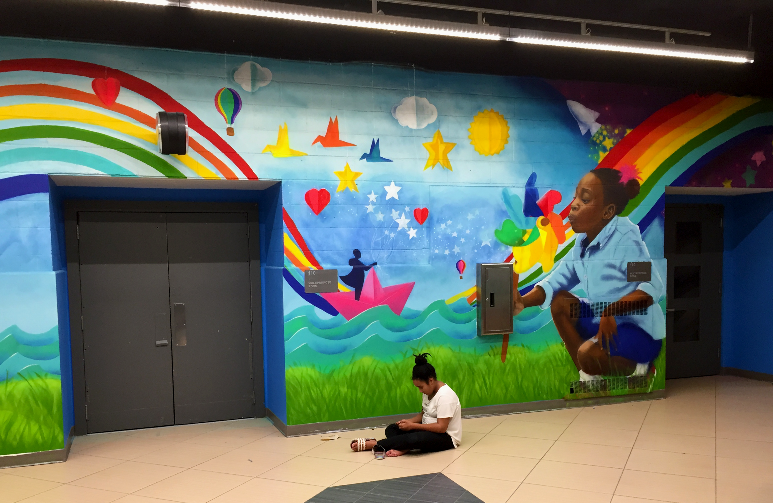 A young woman sitting on the floor in front of a colorful, whimsical mural with rainbows, clouds, stars, hearts, hot air balloons, and a giant girl holding a butterfly, in an indoor setting.