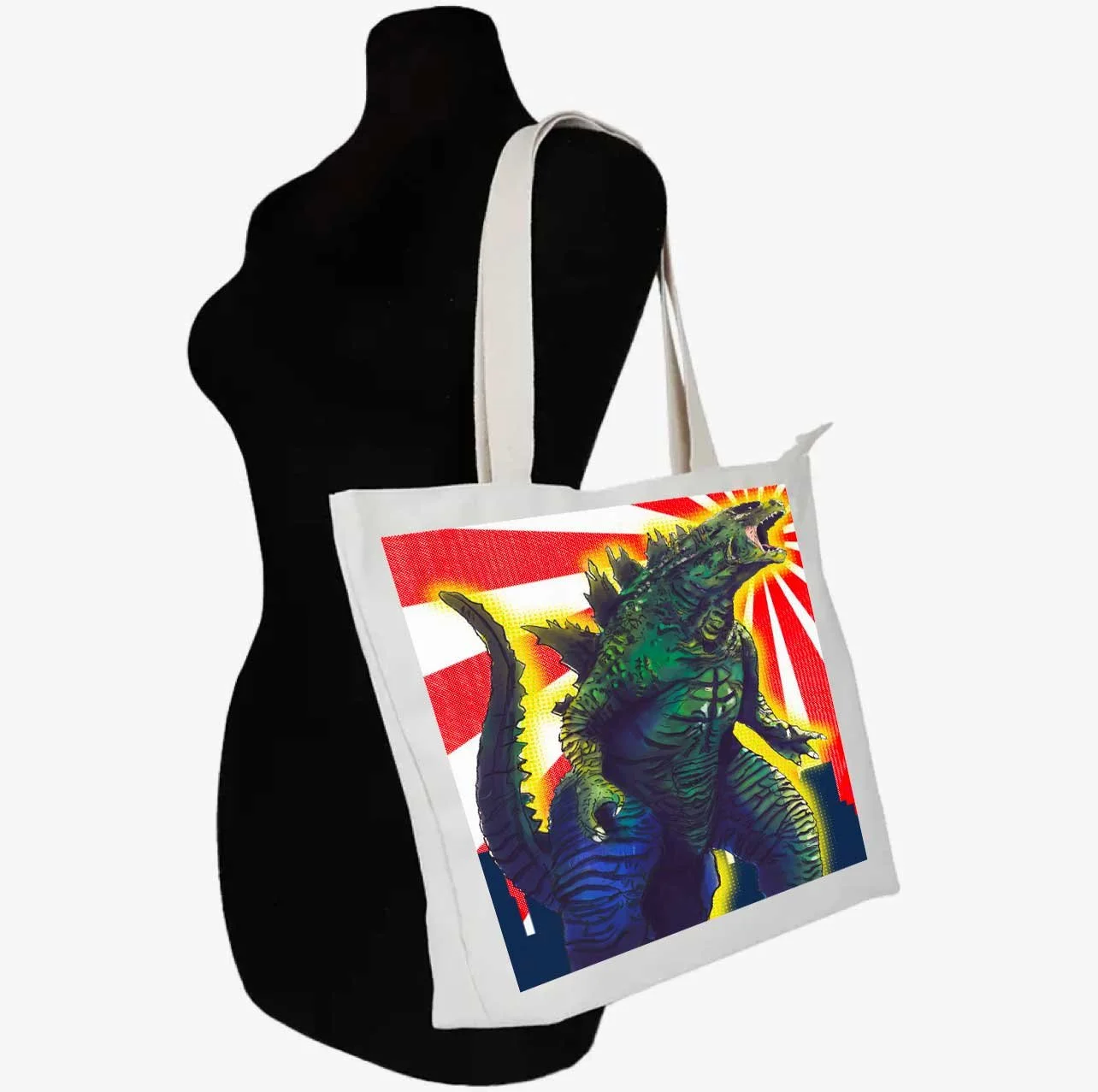 A white tote bag with a colorful graphic of a green Godzilla monster on it, hanging on a black mannequin torso.
