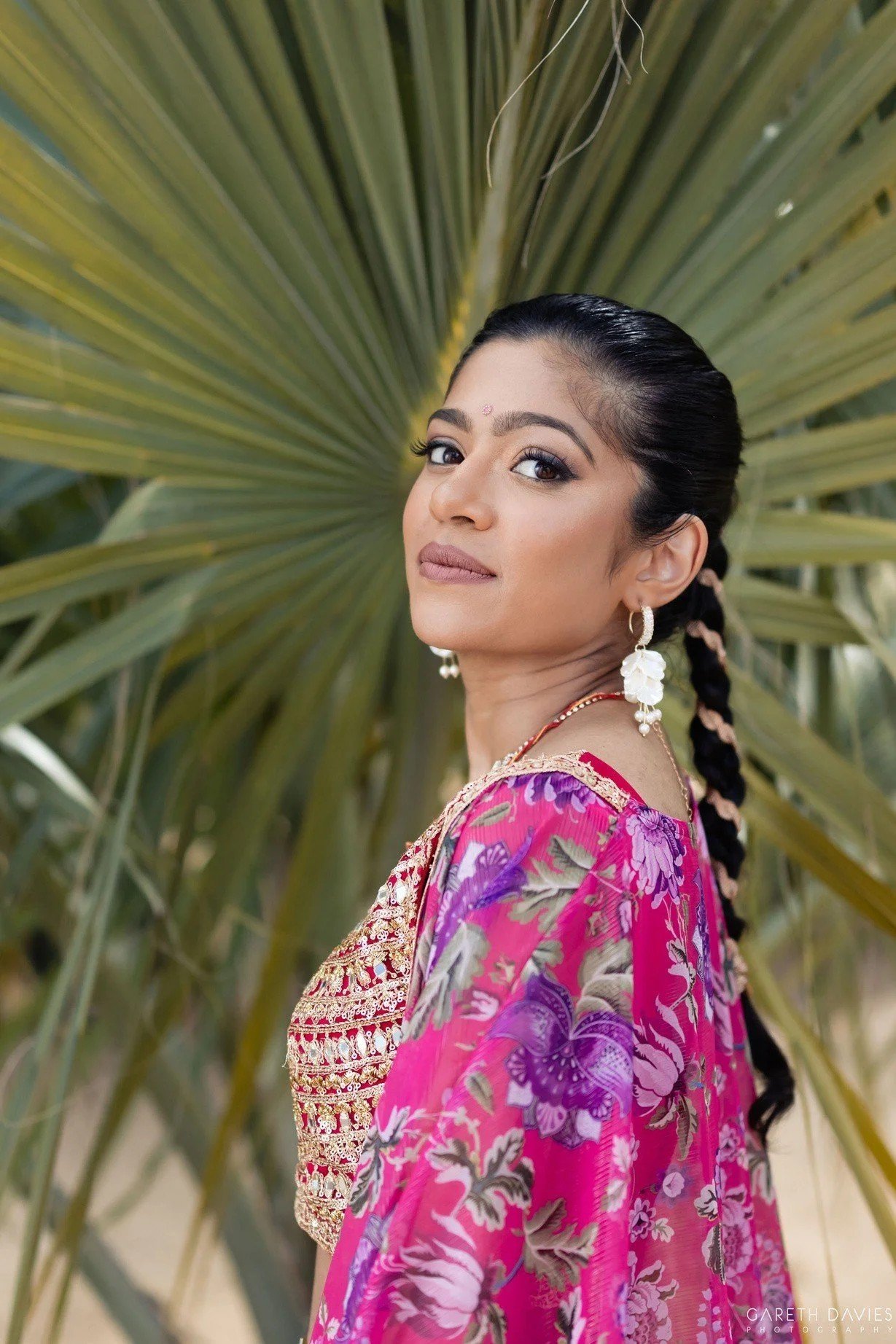 Bride in pink floral lehenga with braided hairstyle among palm leaves, destination wedding by Red Lotus Artistry