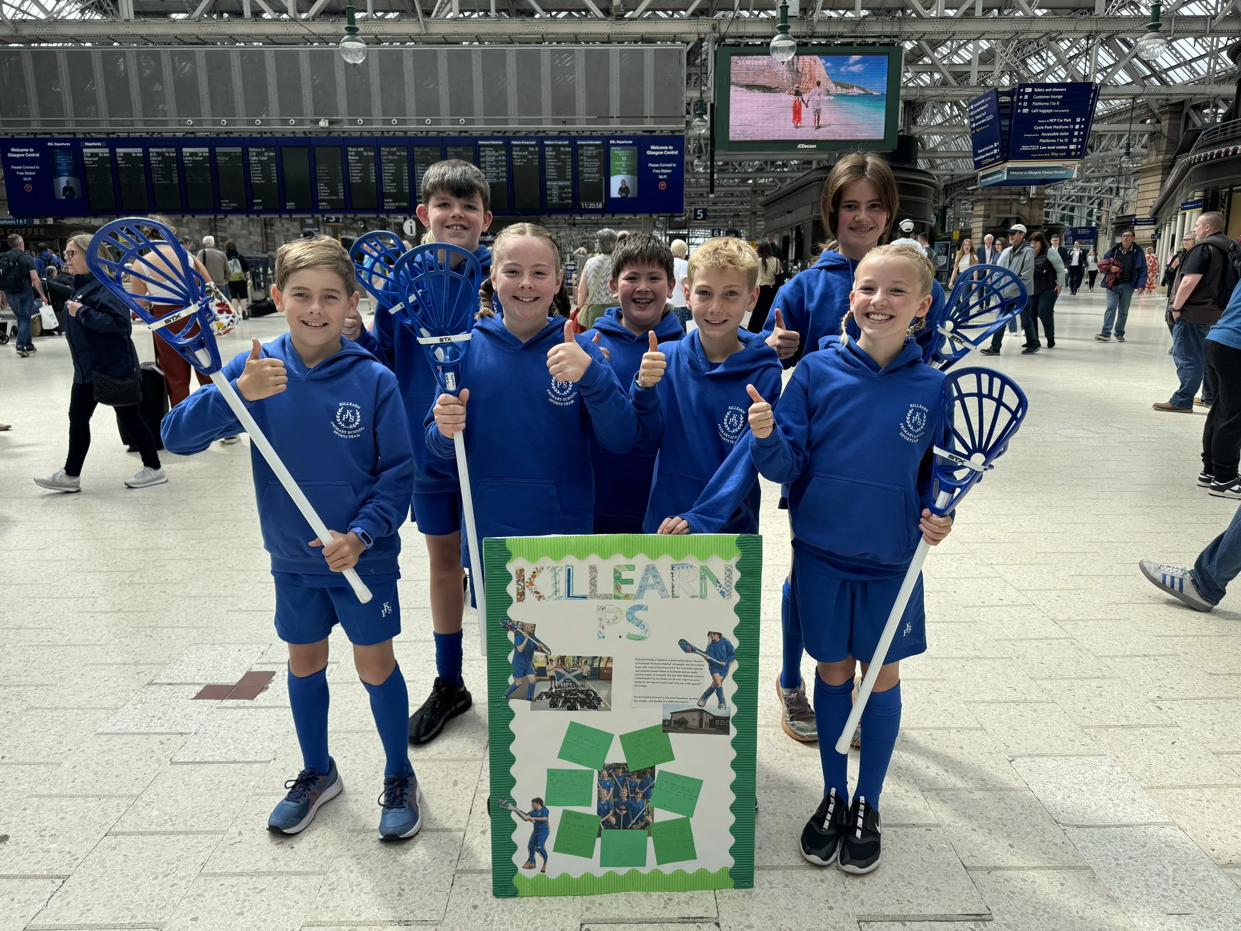 A group of children dressed in blue with pop lacrosse sticks at a railway station. They are all smiling and some have their thumbs up