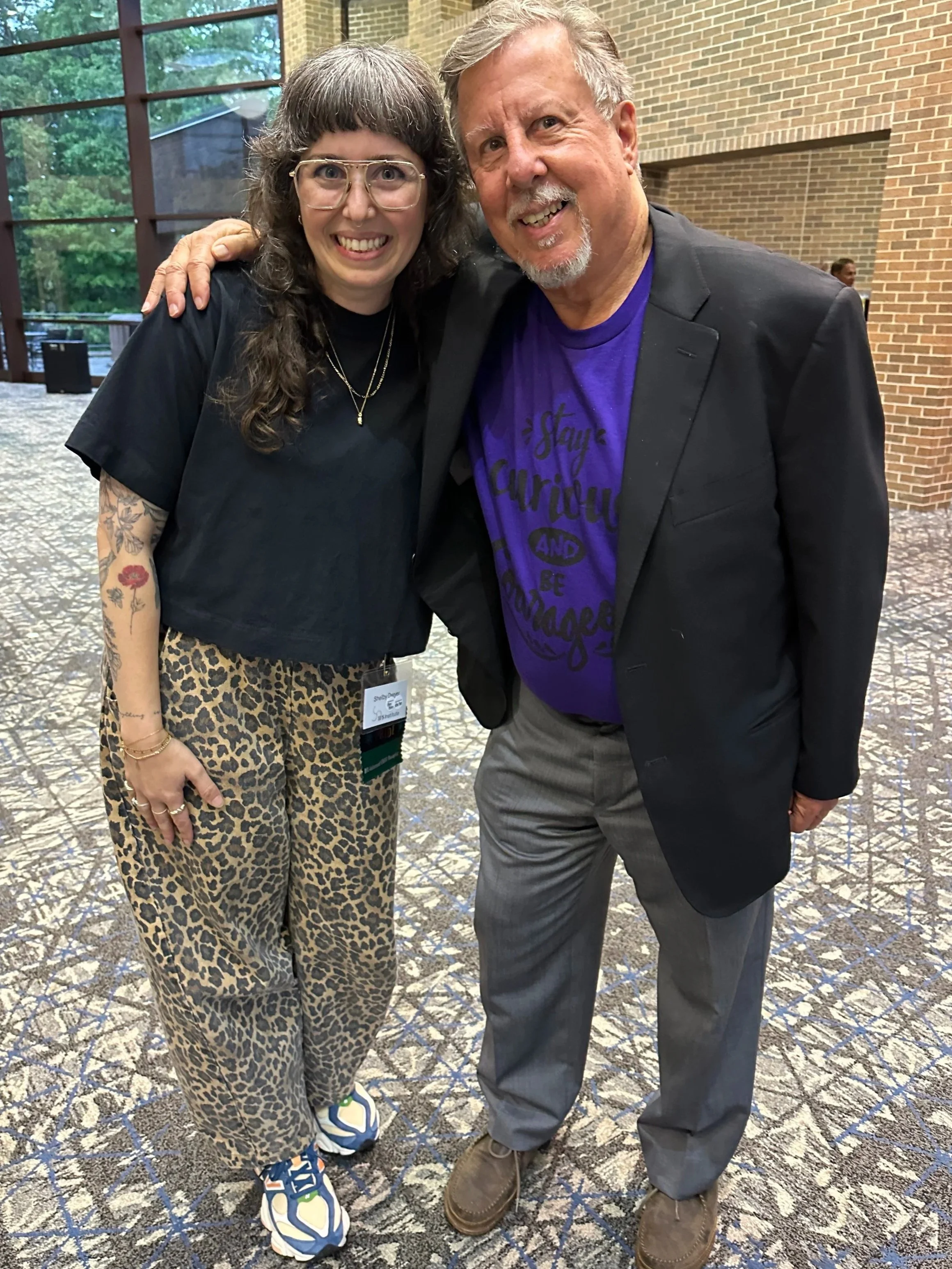 A photograph of a white femme with graying brown hair, gold/silver glasses, wearing a black shirt and leopard-printed pants, with their arm around a man in a black blazer, purple teeshirt, and gray pants.