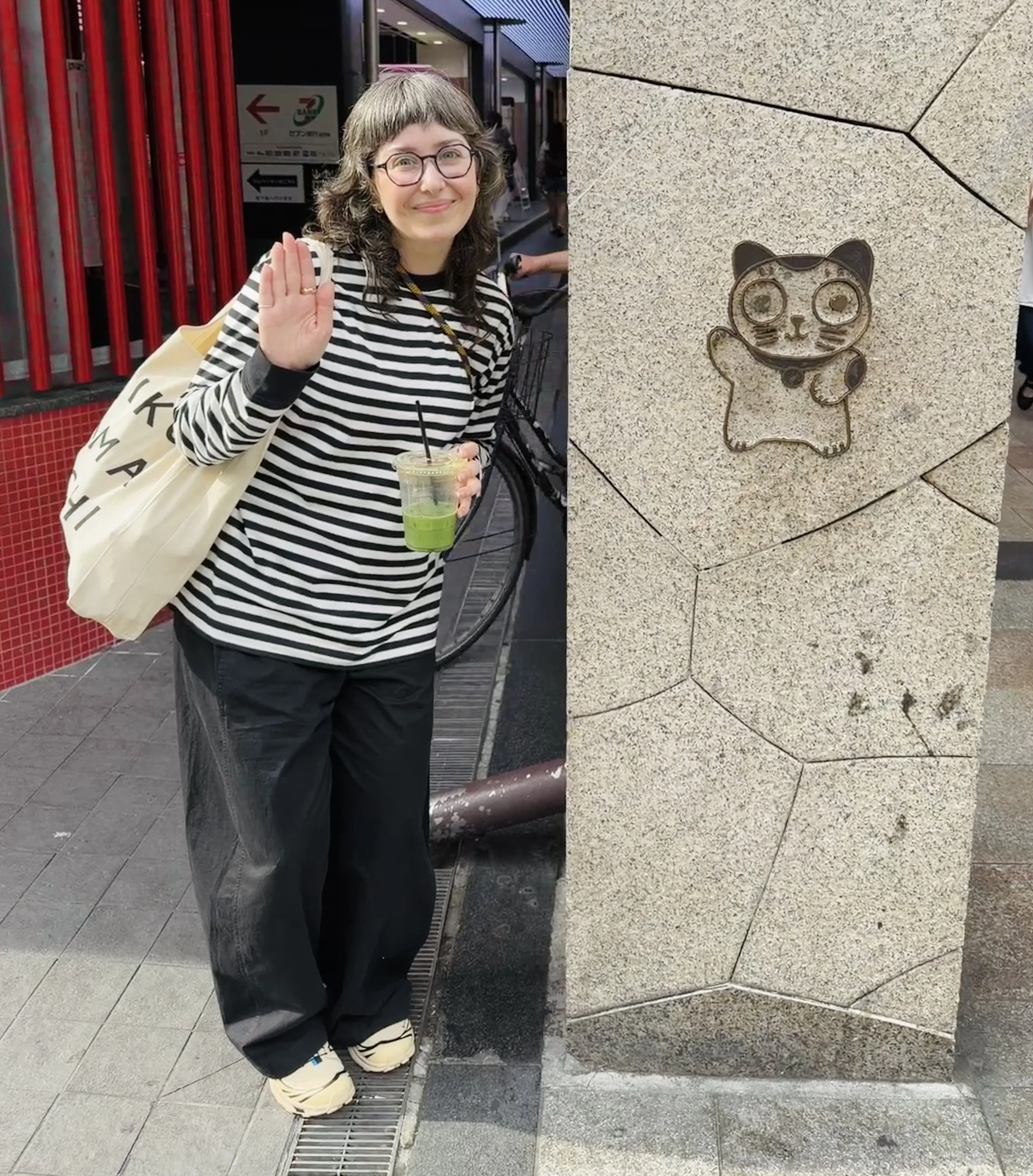 Photo of a white person with graying brown hair, blue glasses, wearing a striped black and white shirt, black pants, and tan sneakers. They are waving hello and standing next to a pillar with an image of a waving cat on it.