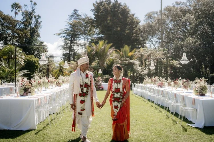 A bride and groom holding hands at an outdoor wedding reception, with long tables decorated with flowers and candles on either side, in a garden setting with trees and palm plants.
