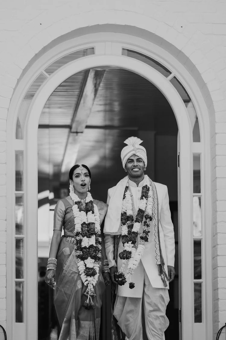 A traditional Indian wedding scene with a bride and groom standing at the entrance of a building, both wearing garlands and traditional attire, with the groom wearing a turban and the bride in a saree.