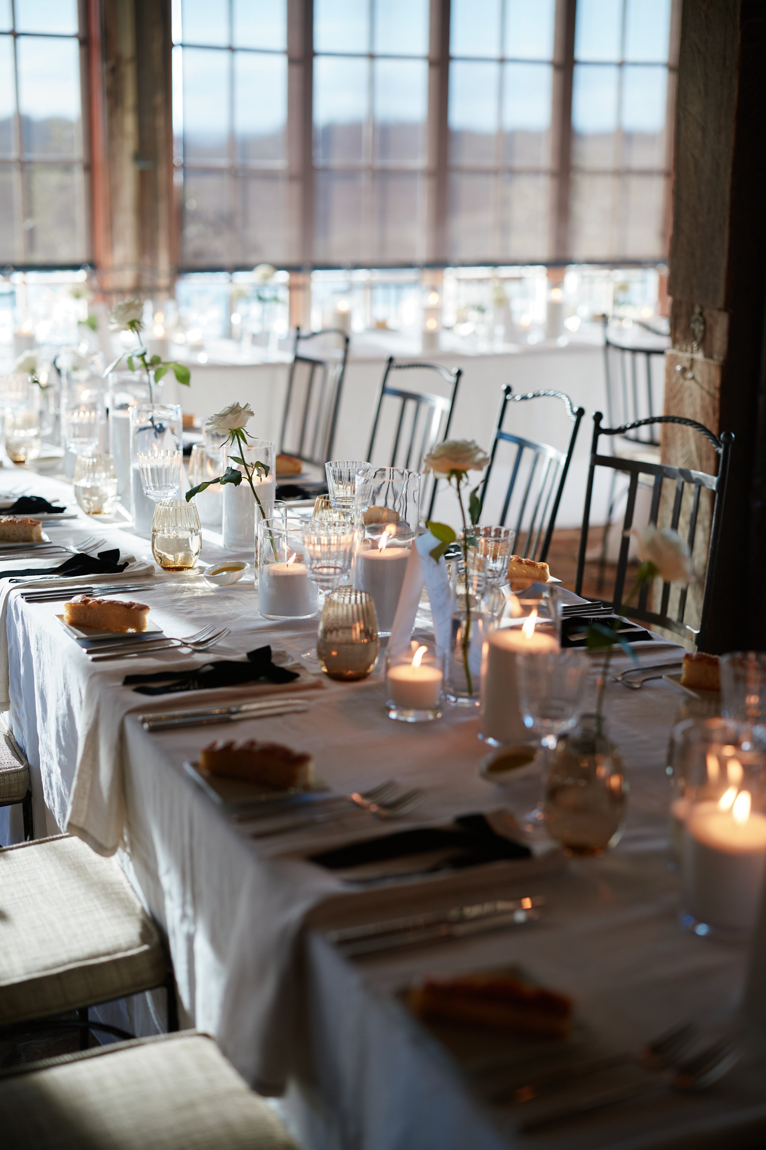Elegant dining table set for a special occasion with candles, white roses in vases, and tableware, near large windows letting in natural light.
