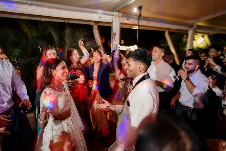 A wedding reception with guests dancing and celebrating, featuring a newlywed couple in traditional attire surrounded by friends and family under a decorated outdoor shelter.