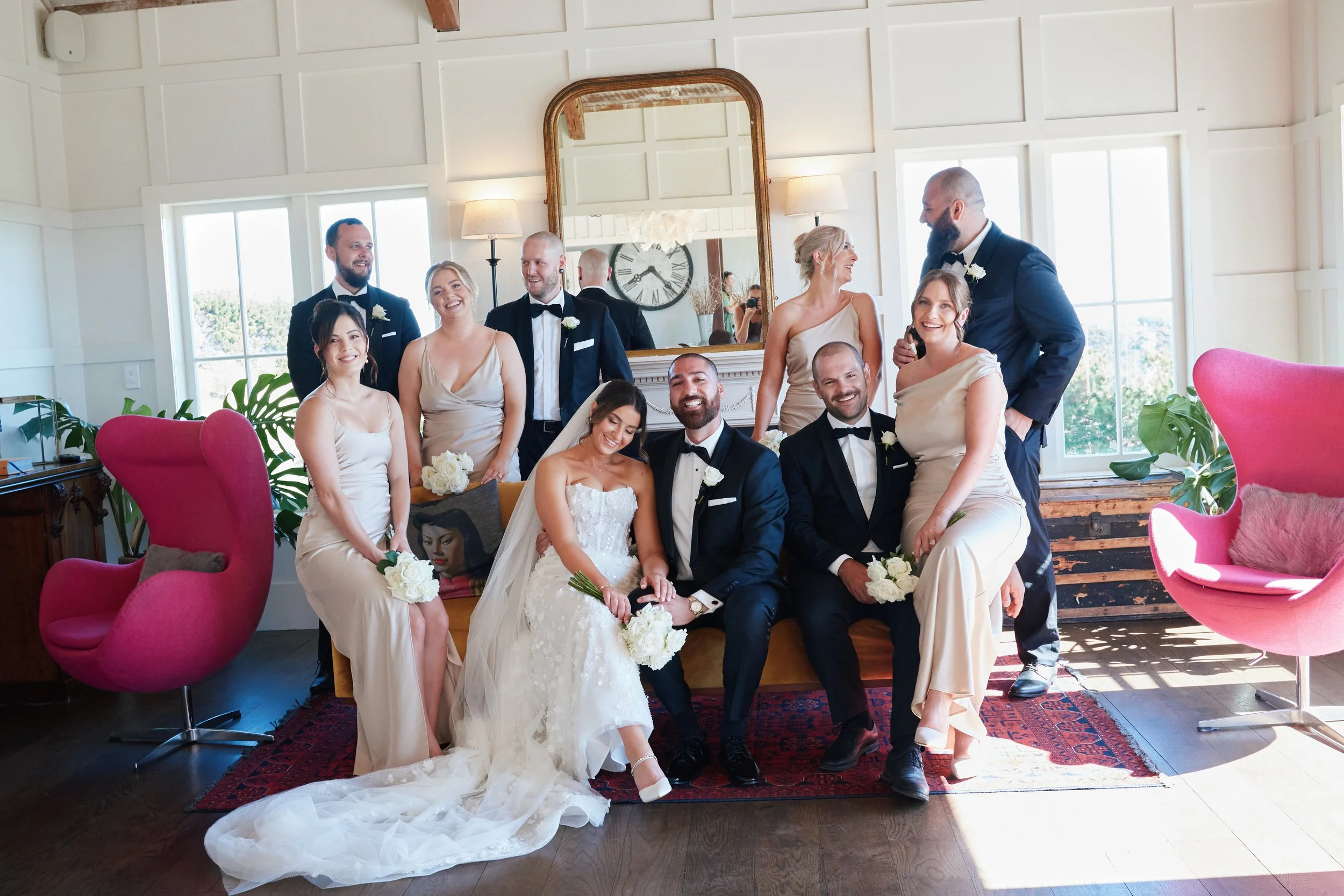A group of wedding guests, including the bride and groom, celebrating indoors near a large window and mirror, with pink chairs and green plants.