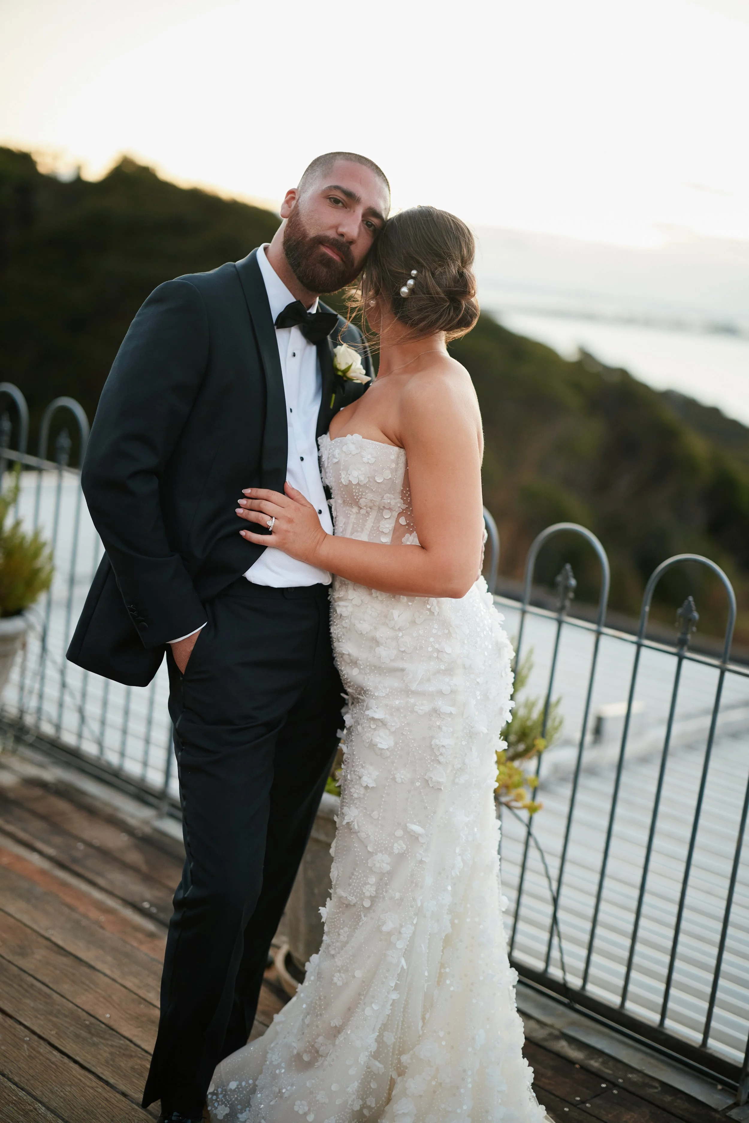 A bride and groom standing on a wooden deck during sunset, with the bride holding the groom's waist and the groom with one hand in his pocket. The groom is wearing a black tuxedo with a bow tie, and the bride is in a strapless white wedding gown with