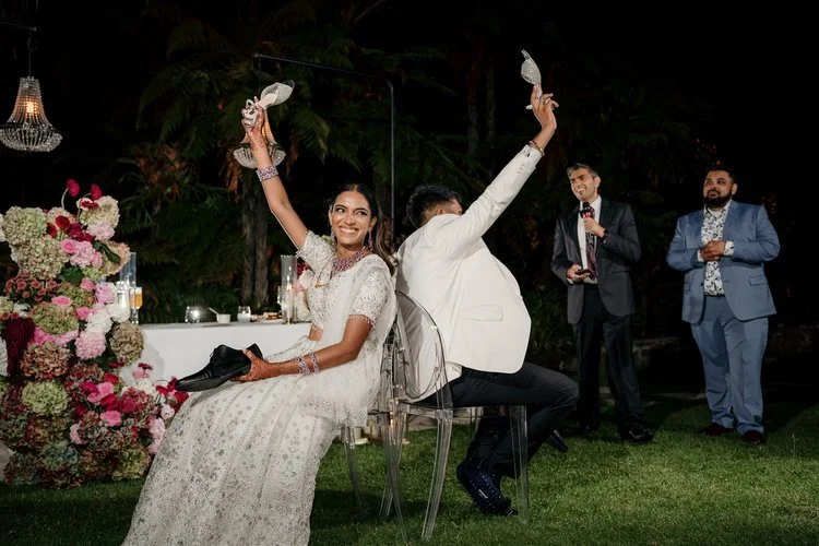 A joyful bride in a white wedding dress holding a shoe and a champagne glass, sitting on a transparent chair at a wedding reception, with a groom in a white blazer and black pants sitting back-to-back with her, both raising their glasses in a toast. 