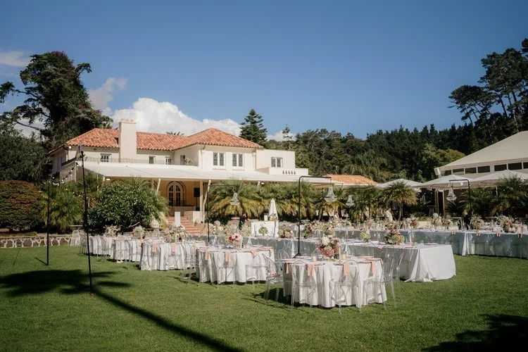 Wedding reception setup with round tables covered in white tablecloths, decorated with floral centerpieces, outdoors on a lush lawn in front of a large house or villa under a clear blue sky.
