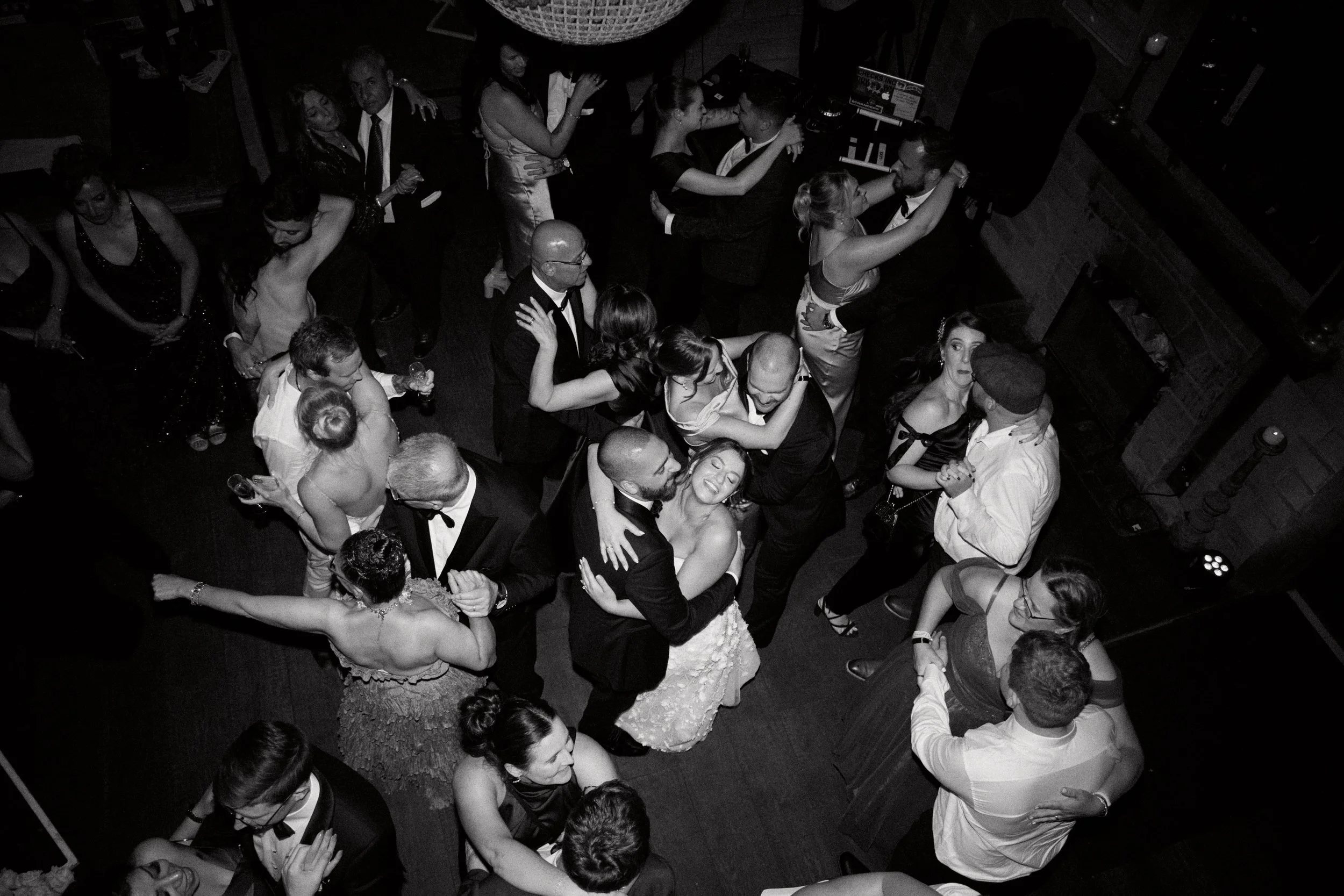 Black-and-white photo of a dance floor at a wedding reception with couples dancing closely together, some in formal attire, in a dimly lit setting with a fireplace in the background.