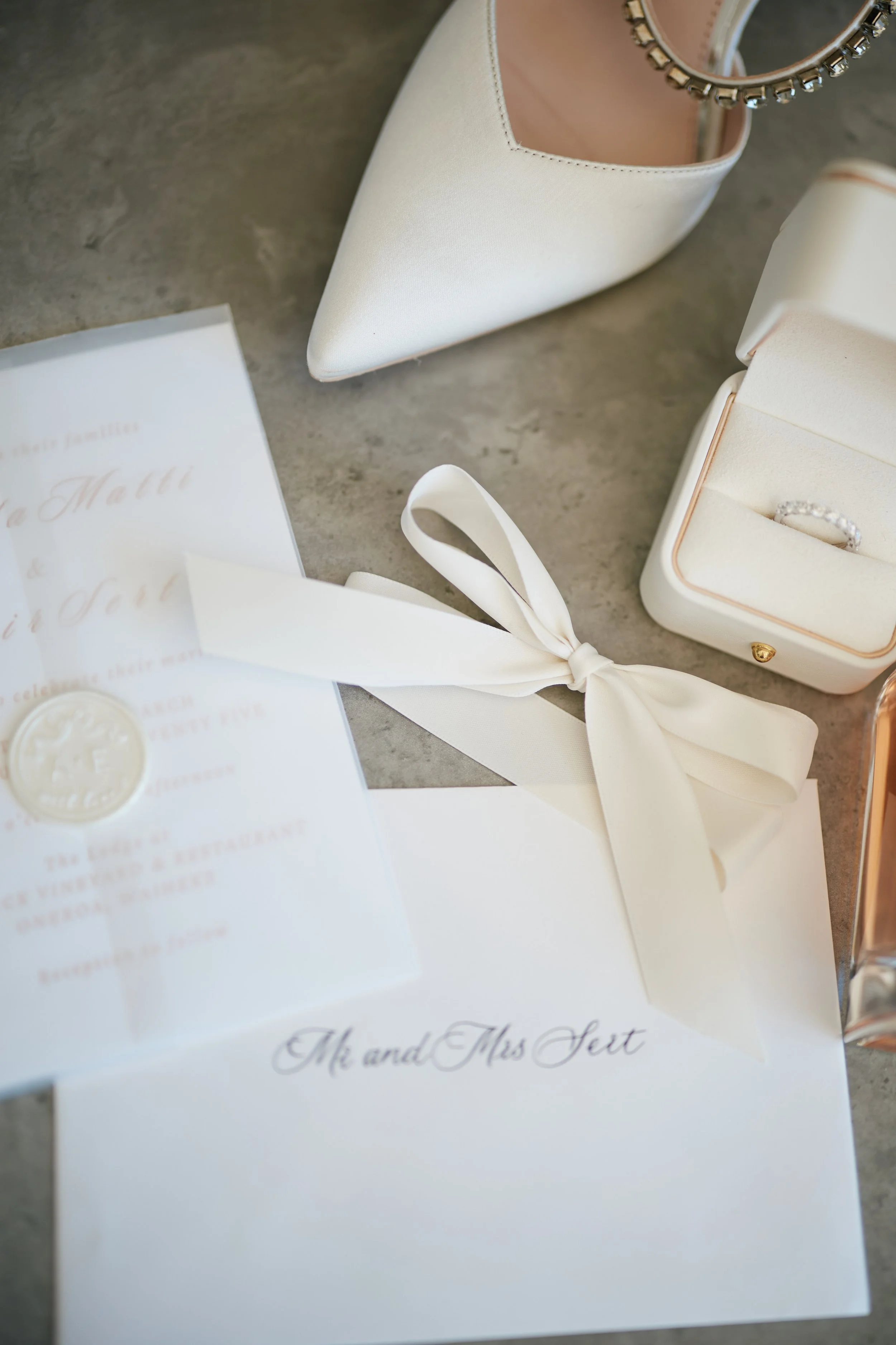 Wedding invitation, white high heel shoe with ankle strap, diamond engagement ring in white box, white ribbon, and gold seal on grey surface.