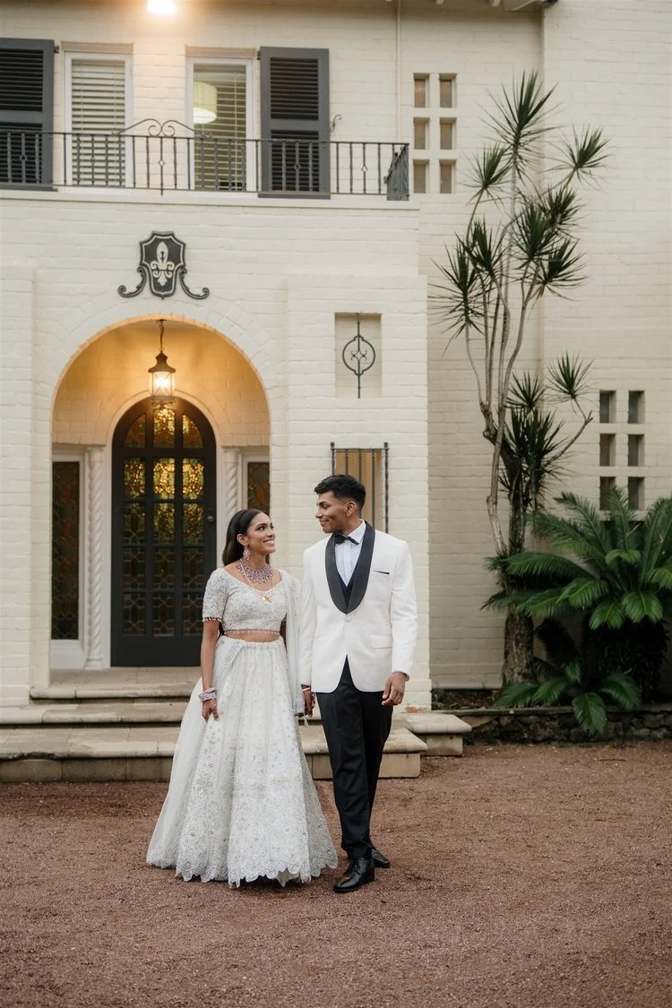 A bride and groom in wedding attire standing outside a light-colored brick house with a large arched doorway and black shutters, surrounded by greenery.