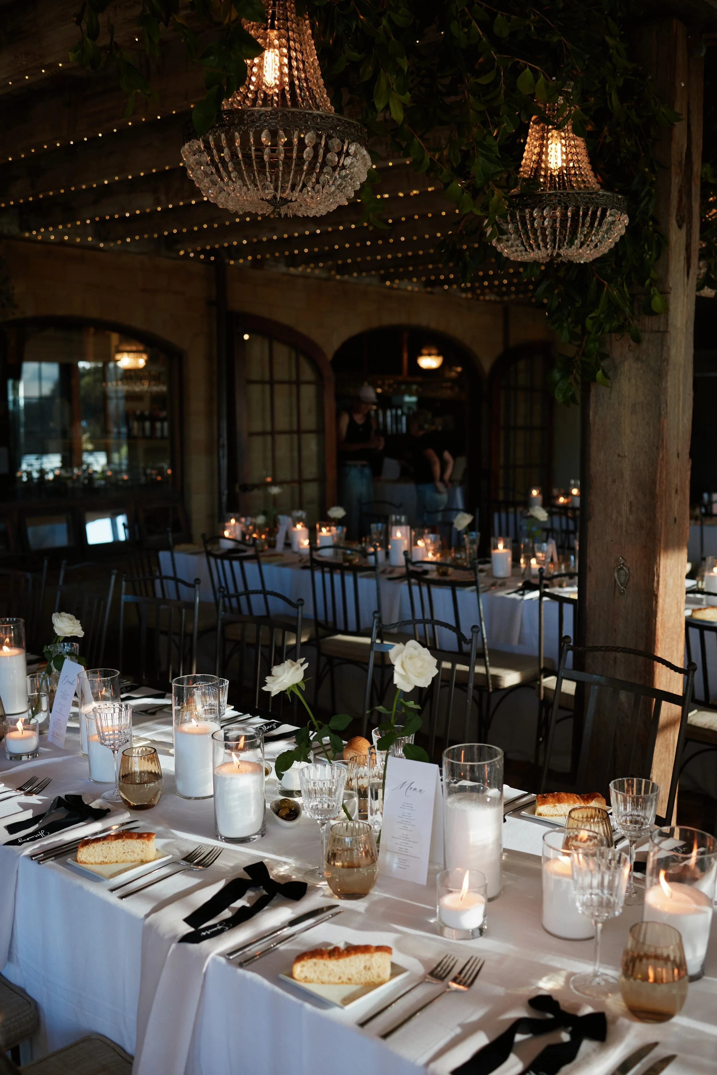 Elegant indoor dining setup with white tablecloths, candles, floral centerpieces, and place settings, under chandeliers and string lights in a rustic restaurant or event space.