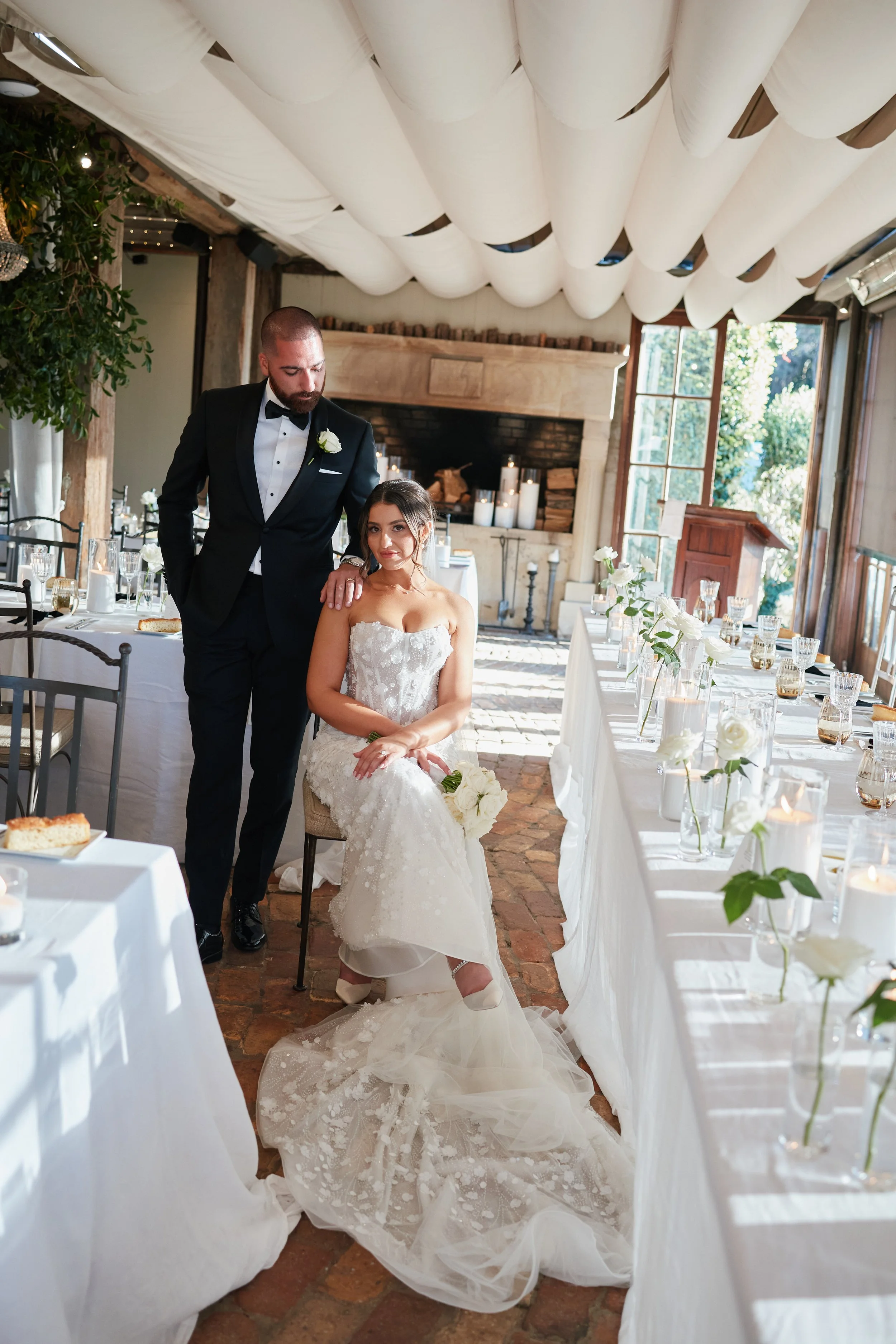 A bride and groom in a decorated wedding reception venue with long banquet tables, white flowers, and candles.