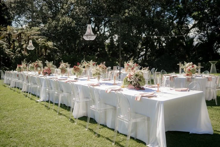 Outdoor wedding reception setup with long tables, white tablecloths, and floral centerpieces, set on a grassy lawn with string lights hanging overhead.
