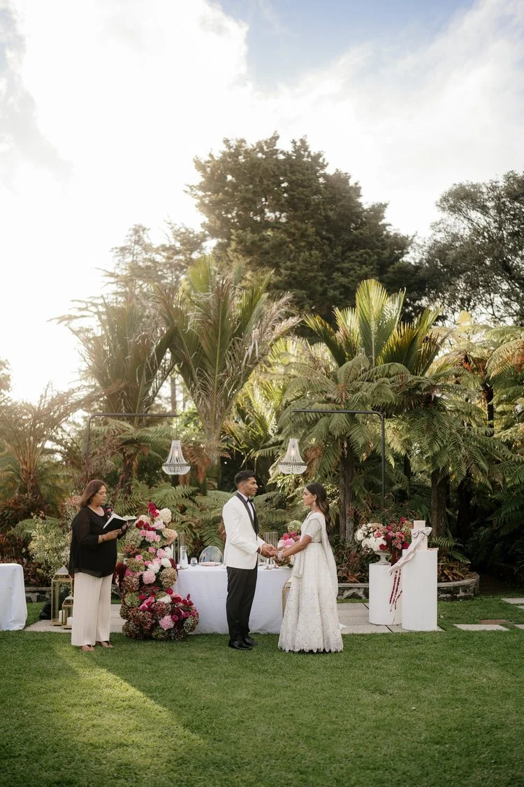 A couple at their wedding ceremony exchanging vows outdoors on a grassy lawn, with a woman officiating and lush green trees in the background.