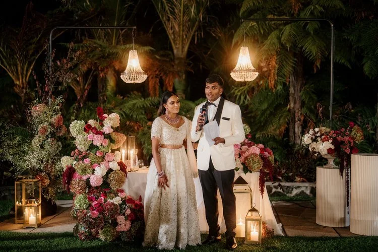 A couple giving a wedding toast outdoors at night, surrounded by flowers, candles, and hanging chandeliers.