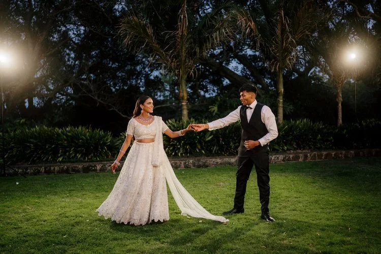 A couple dancing outdoors on a grassy area during dusk, with palm trees in the background and two bright lights shining from the sides.