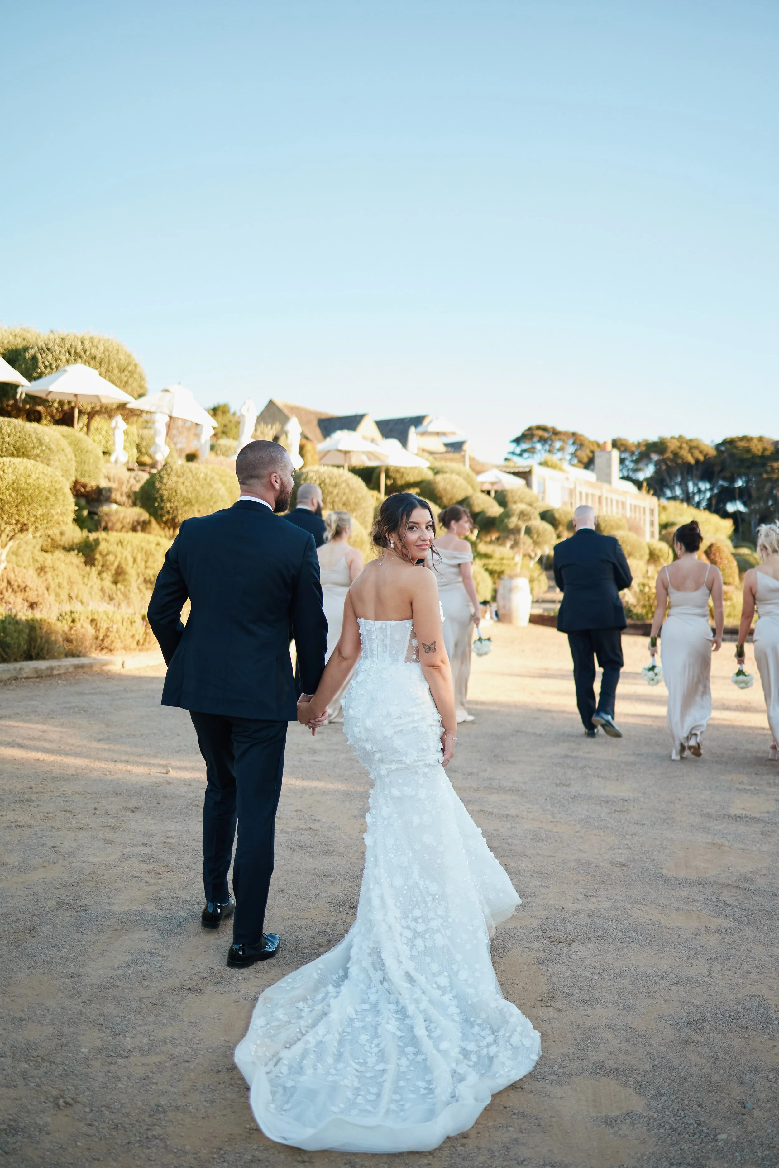 A bride and groom holding hands, walking outdoors on a wedding day, with bridesmaids and groomsmen in the background and a clear blue sky.