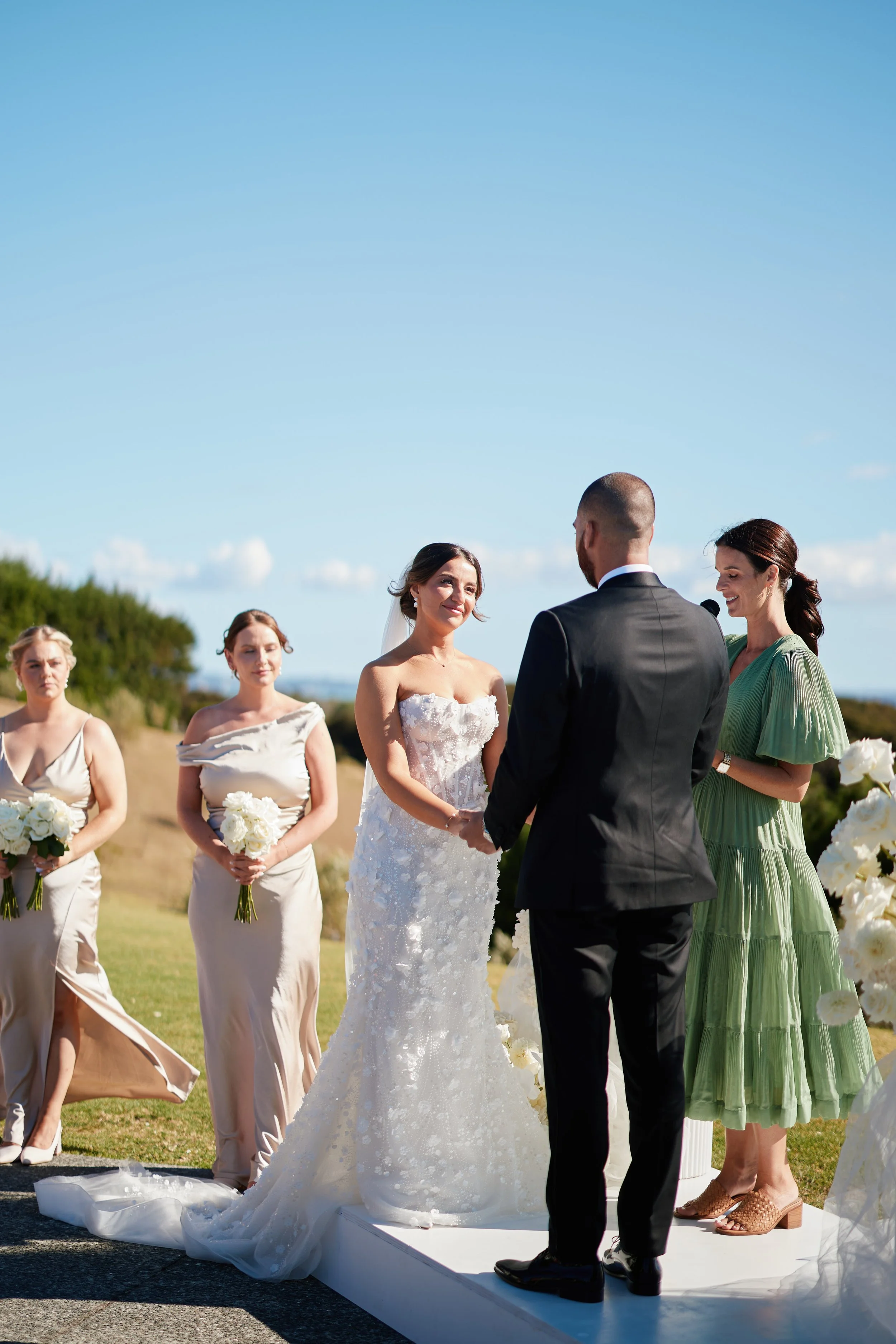A bride and groom exchange vows during an outdoor wedding ceremony with bridesmaids and officiant present under a clear blue sky.