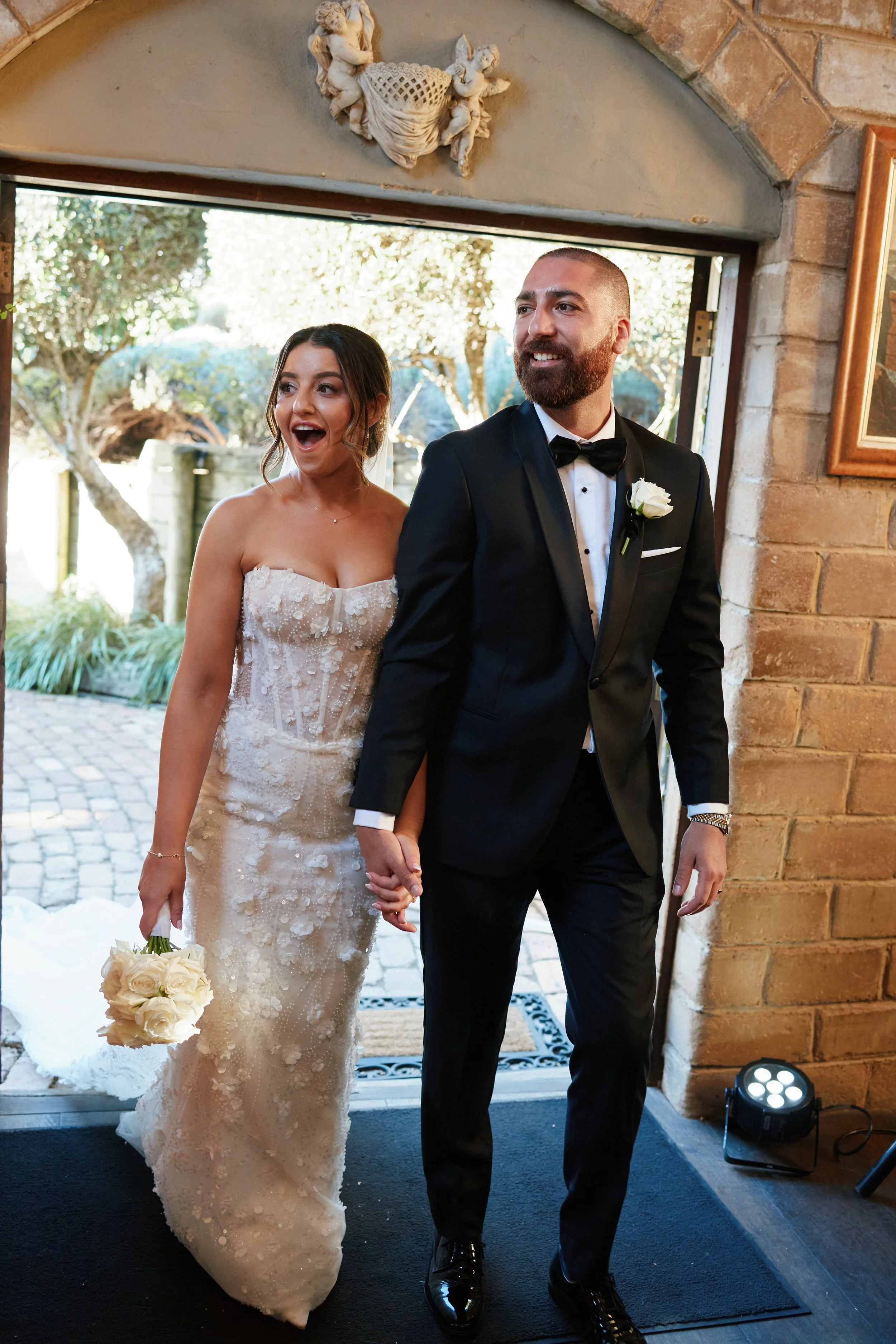 A bride and groom holding hands, walking out of a building at their wedding, with the bride holding a bouquet of white roses and the groom wearing a black tuxedo with a white boutonniere.
