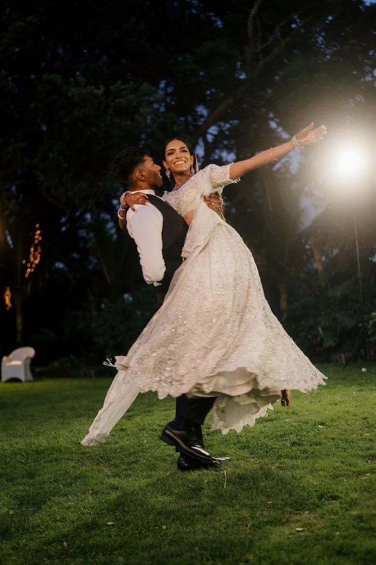 A newlywed couple dancing outdoors at night, with the groom lifting and spinning the bride in a white wedding gown, illuminated by bright lights amidst trees.