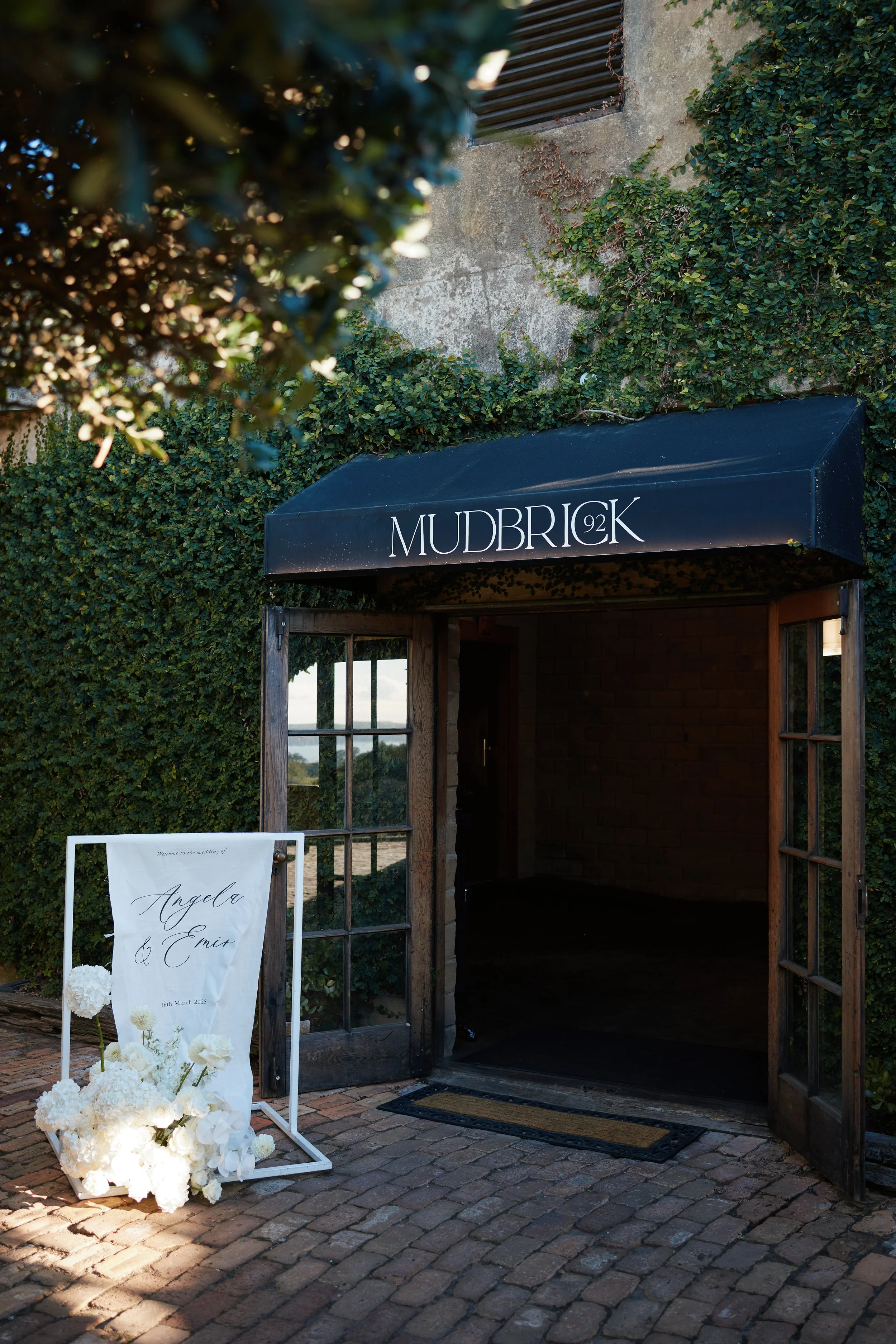Wedding venue entrance with a black awning labeled 'MUDBRICK 92K', open wooden doors, a white sign with black cursive writing announcing the wedding of Angela and Emmet, dated 16th March 2025, decorated with white flowers, and surrounded by green ivy