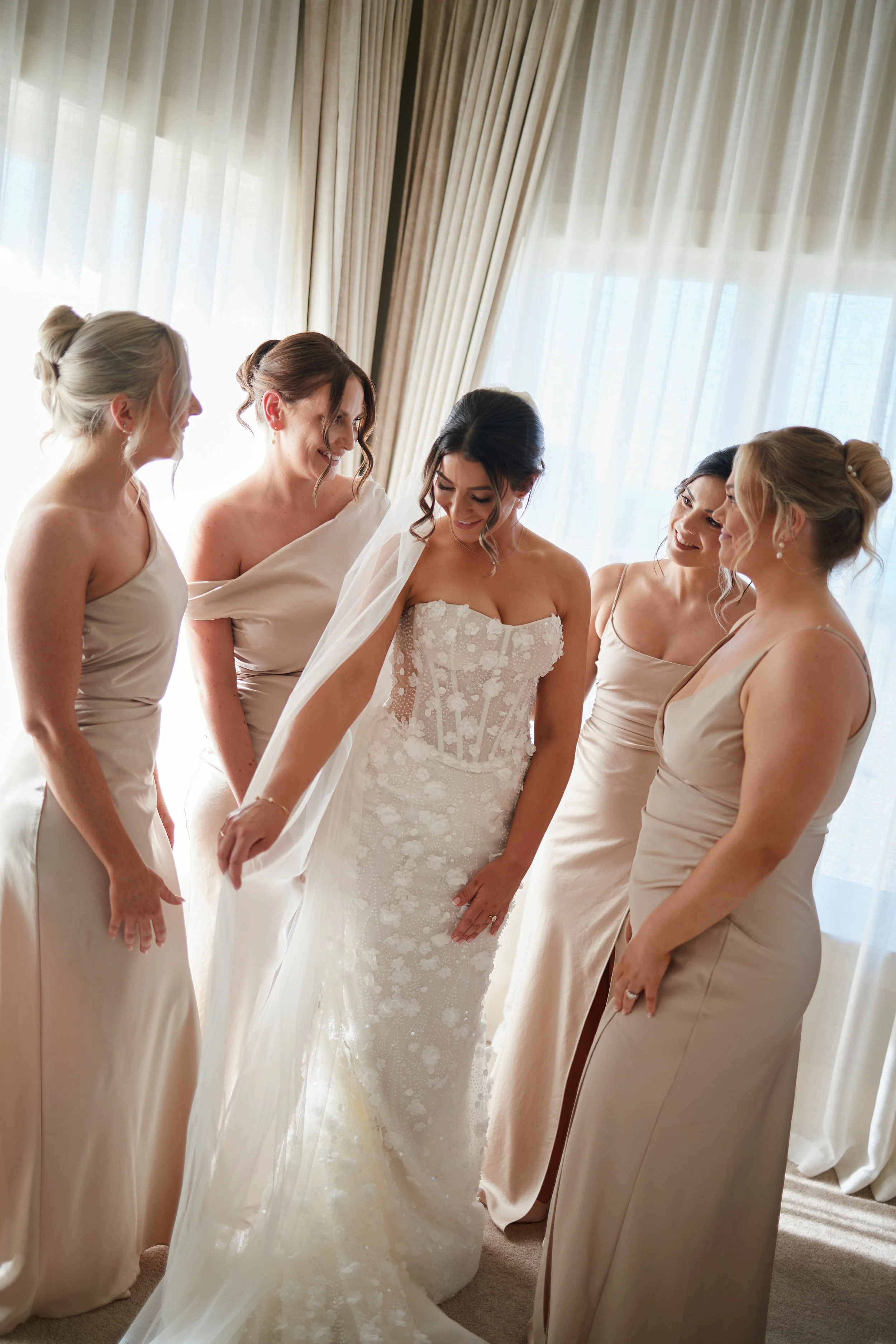 Bride in a white wedding gown with her bridesmaids in beige dresses, all smiling and gathered near a window with sheer curtains.