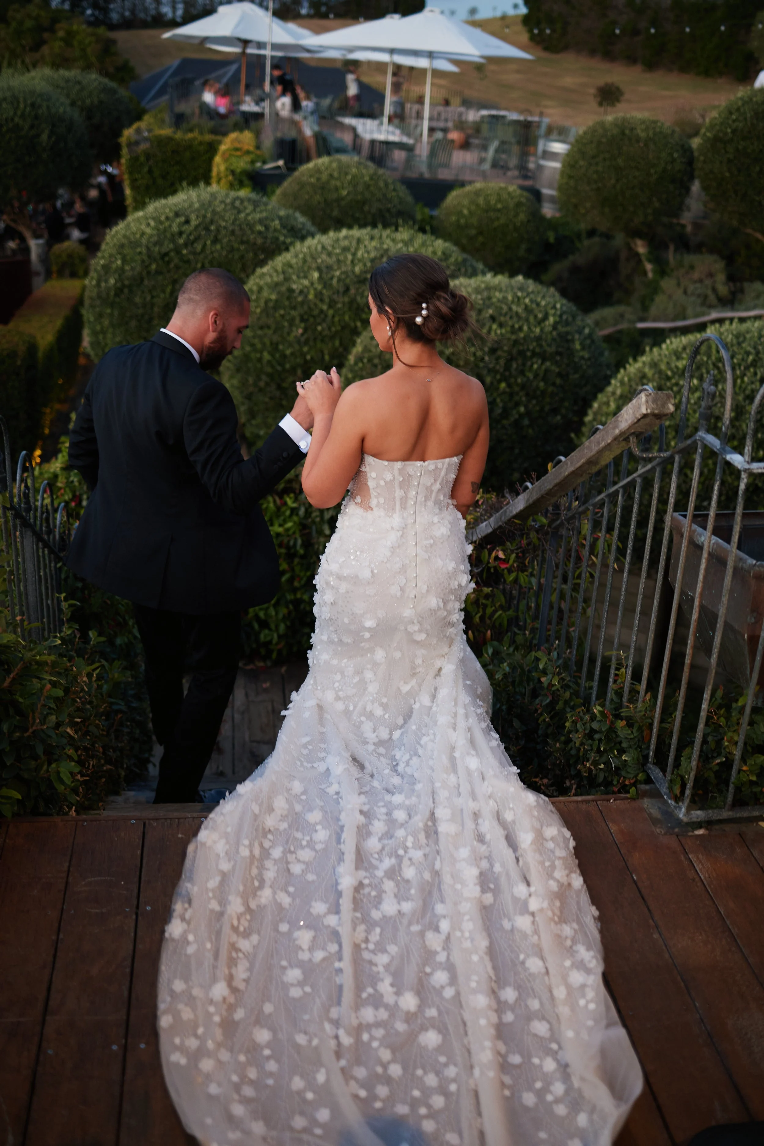 A bride and groom holding hands on a staircase outdoors at sunset, with the bride wearing a strapless white wedding gown with floral appliques and the groom in a black tuxedo, surrounded by greenery and other wedding guests in the background.