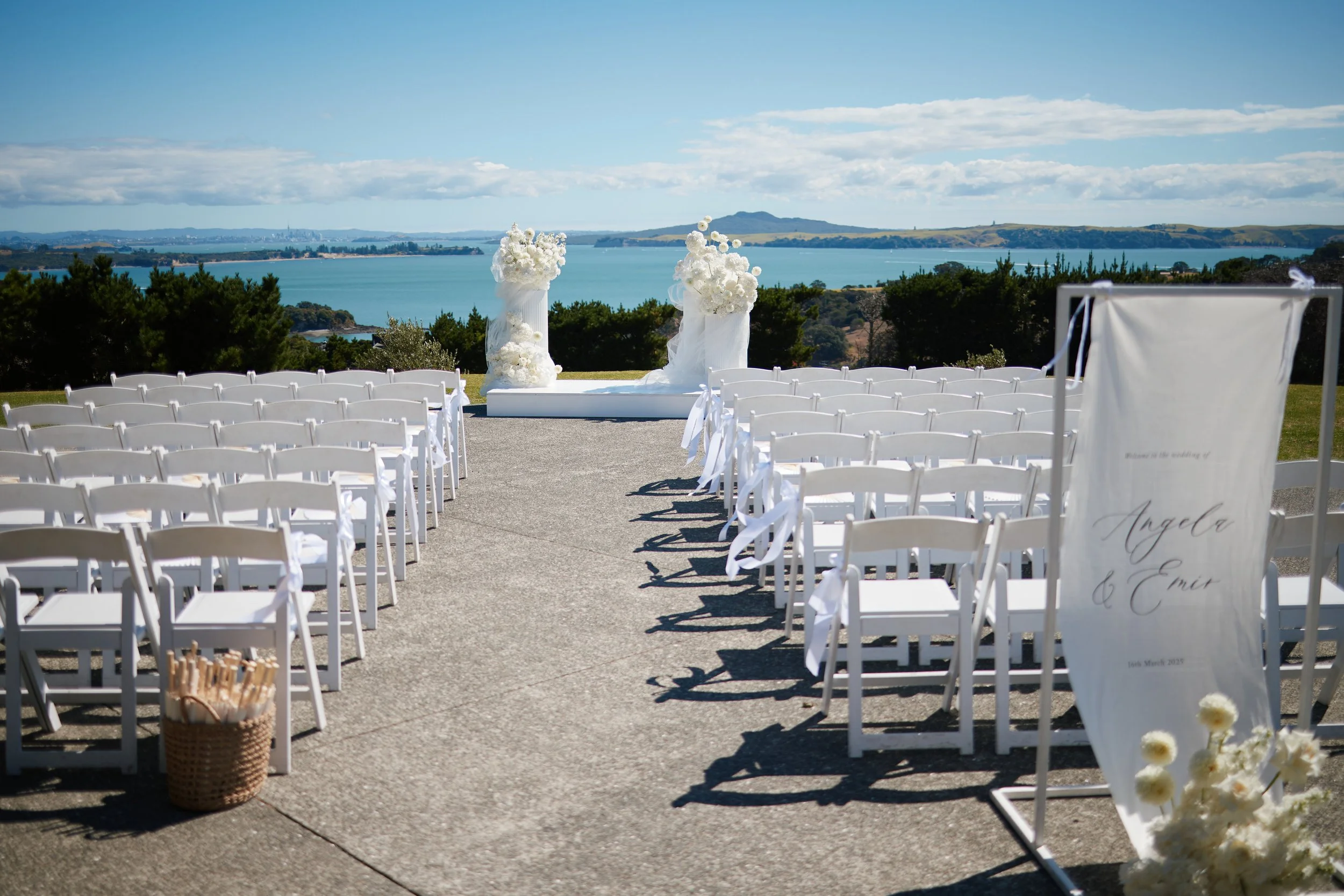 Outdoor wedding ceremony setup with rows of white chairs facing an altar with flowers, overlooking a body of water and hills under a blue sky.