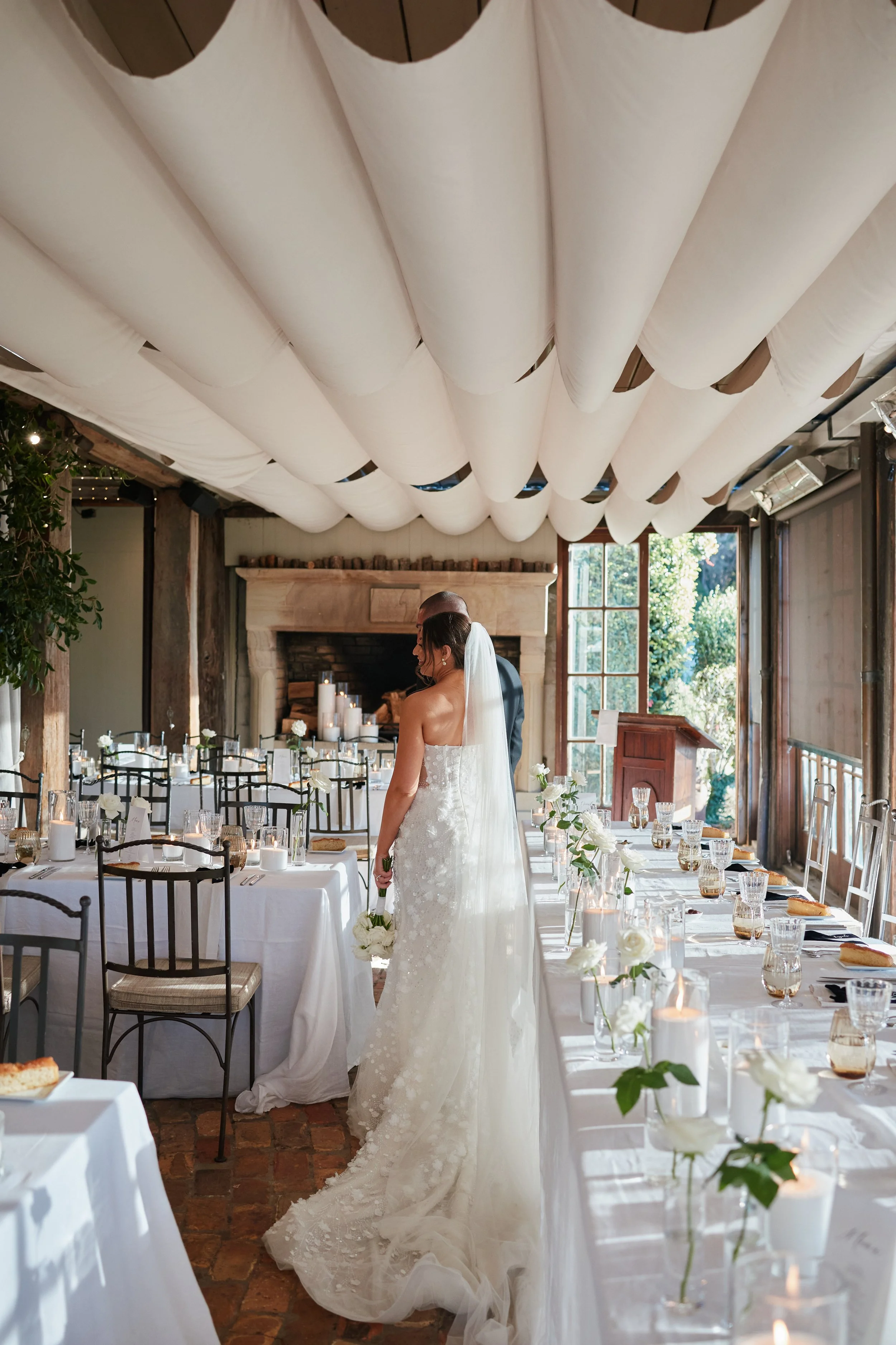 Bride in a white wedding gown and veil standing next to a man in a tuxedo in a decorated reception hall with a fireplace, candles, roses, and tables set for a wedding celebration.