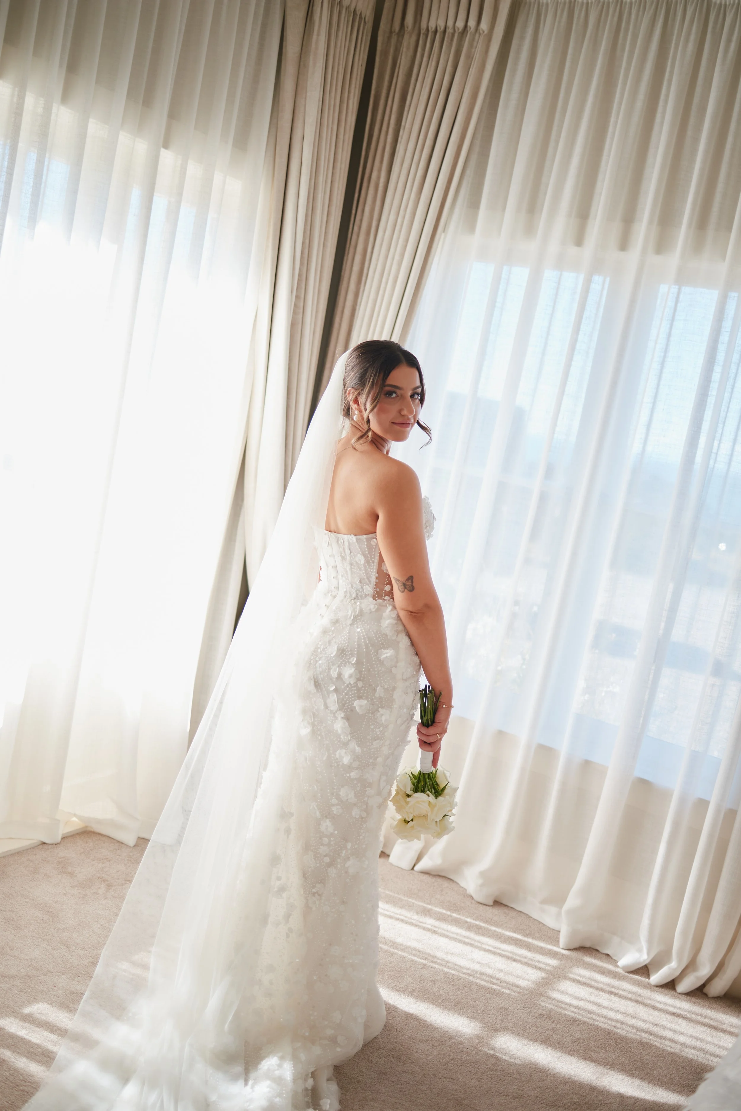 A bride in a white lace wedding gown and veil, holding a bouquet of white flowers, standing indoors near window with sheer curtains.