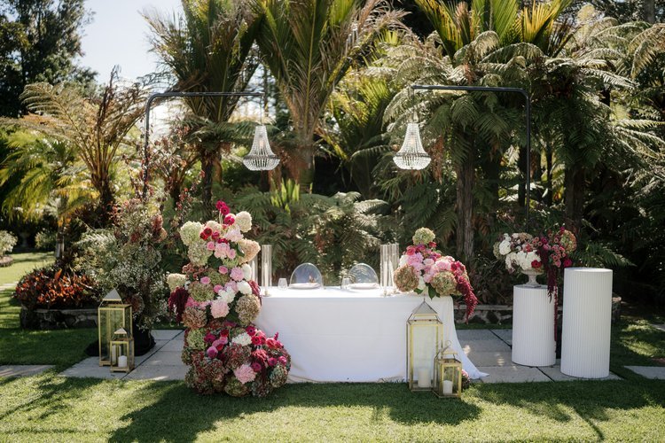 A decorated outdoor garden setting with a white table, floral arrangements, lanterns, and hanging chandeliers for Ahranni and Kritesh at Puketutu.