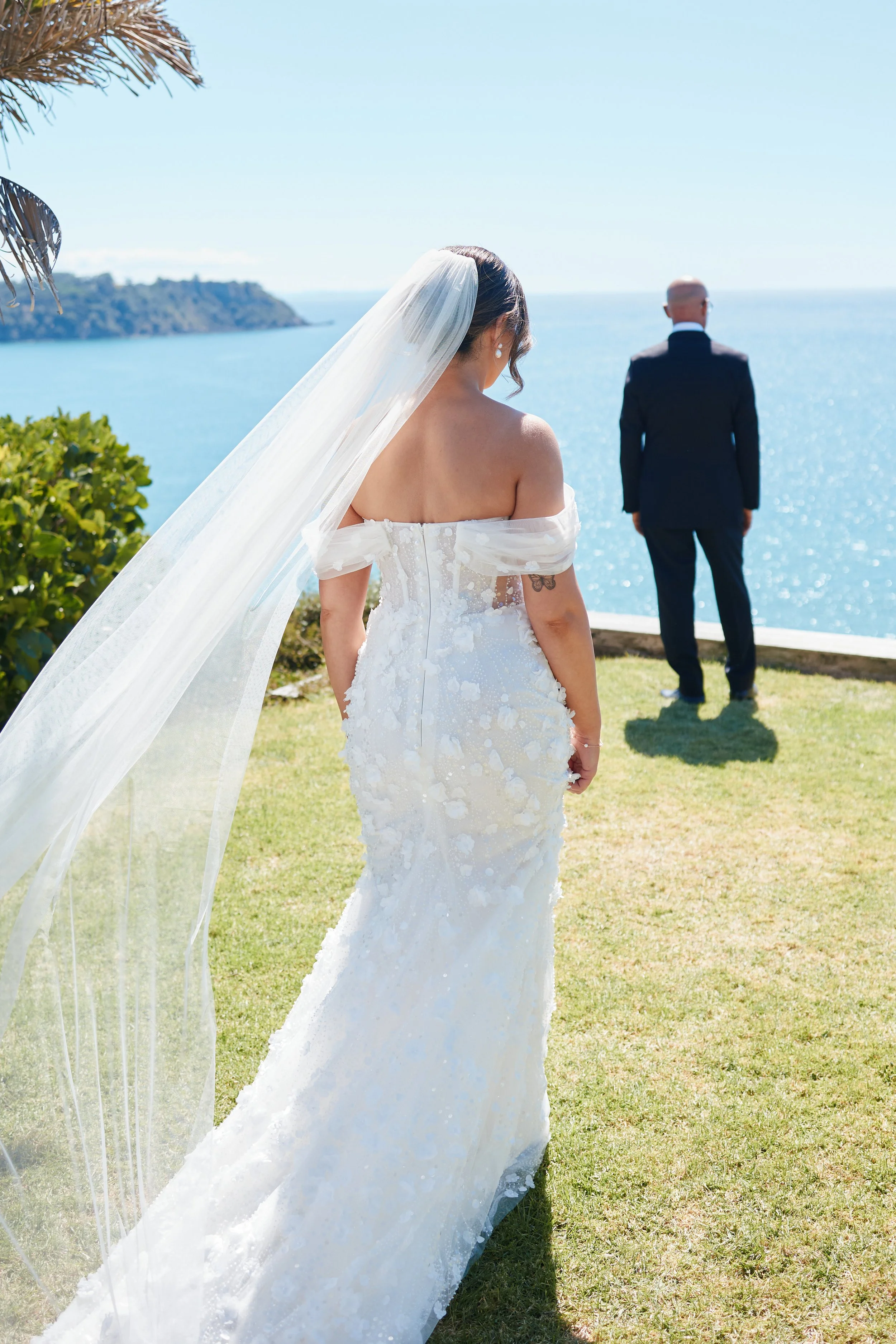 A bride in a white wedding dress with a long veil stands on a grassy area, looking out over the ocean towards a groom dressed in a dark suit who is facing away from her, with a clear blue sky and water in the background.