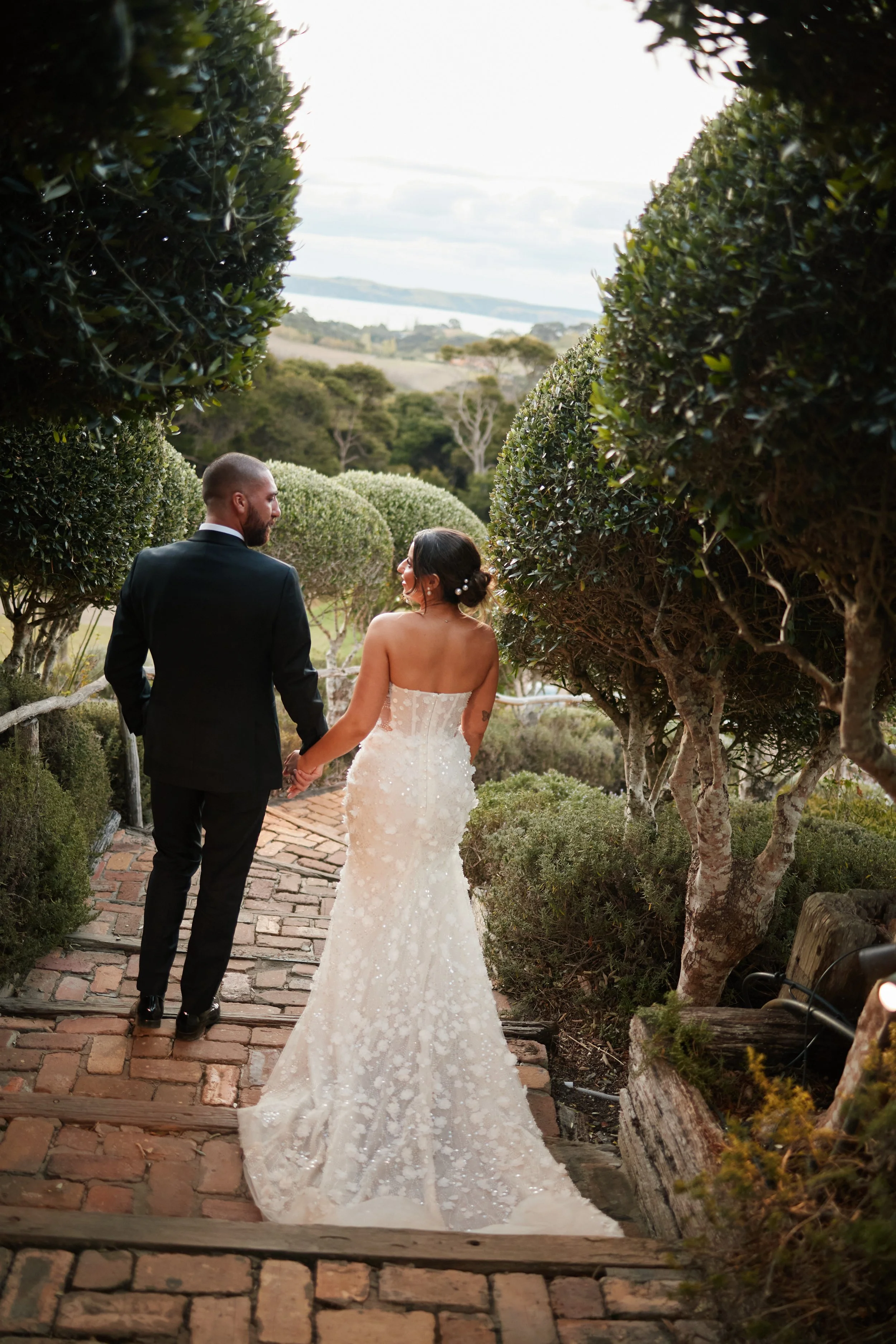 A bride and groom walking hand in hand along a brick pathway through a garden with trees, overlooking a scenic landscape with water and fields in the distance.