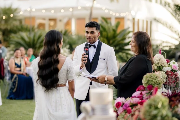 A wedding ceremony outdoors with a groom in a white tuxedo, a bride in a white dress, and a woman officiant holding a book. There are guests in the background and colorful flowers in the foreground.