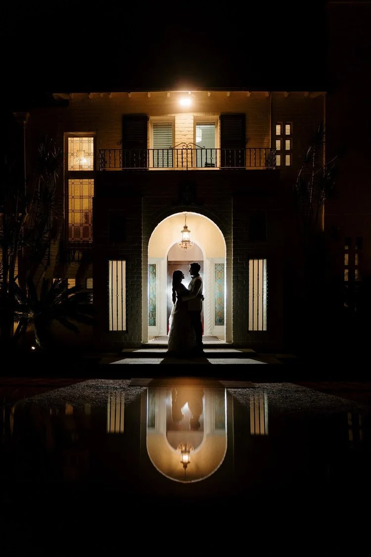 Couple standing in silhouette under an archway, reflected in a puddle outside a warmly lit house at night.