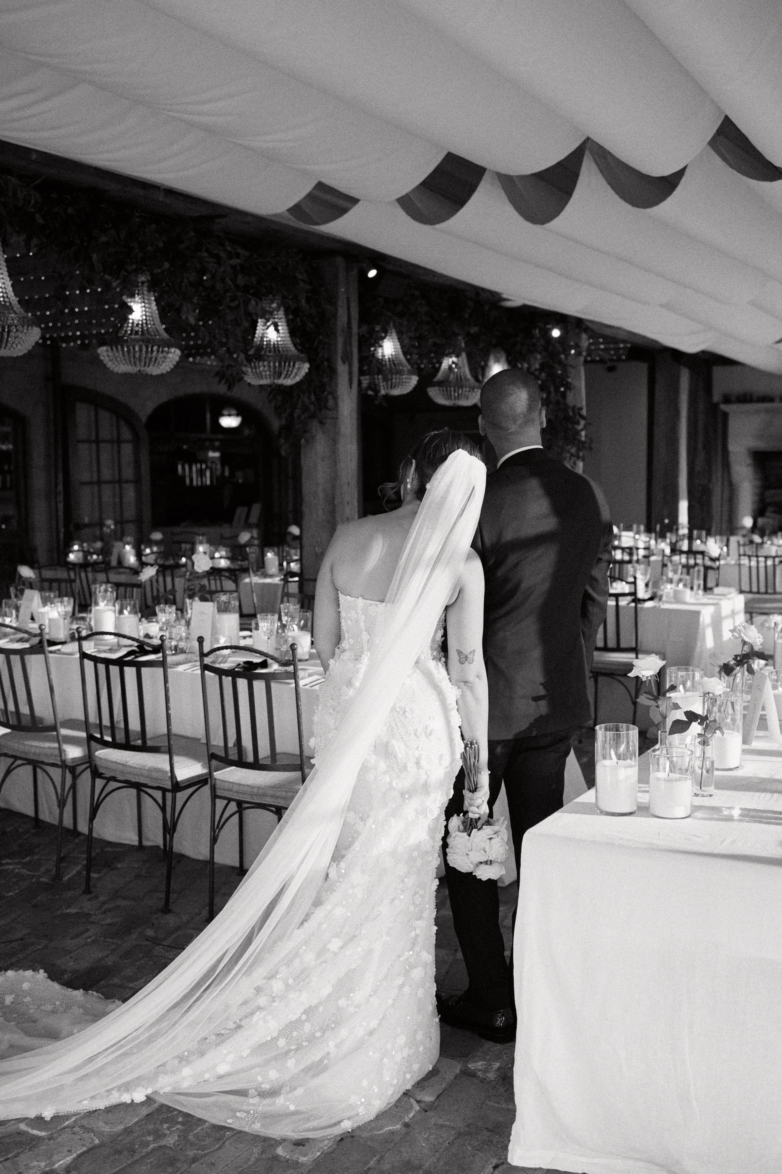 A newlywed couple, a bride in a wedding dress with a long veil and a groom in a suit, walking hand-in-hand at their wedding reception in a decorated hall with tables, chairs, and elegant lighting.