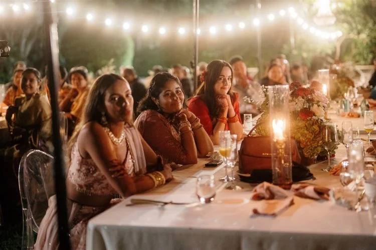 People sitting at a decorated outdoor event table during the evening, with string lights overhead and floral centerpieces.