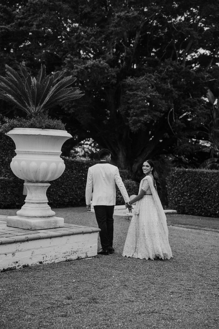 A black and white photo of a couple holding hands, walking outdoors near a large decorative planter and a hedge, with a tall tree in the background. The woman is smiling and wearing a lace dress, the man is dressed in a white blazer and dark pants.