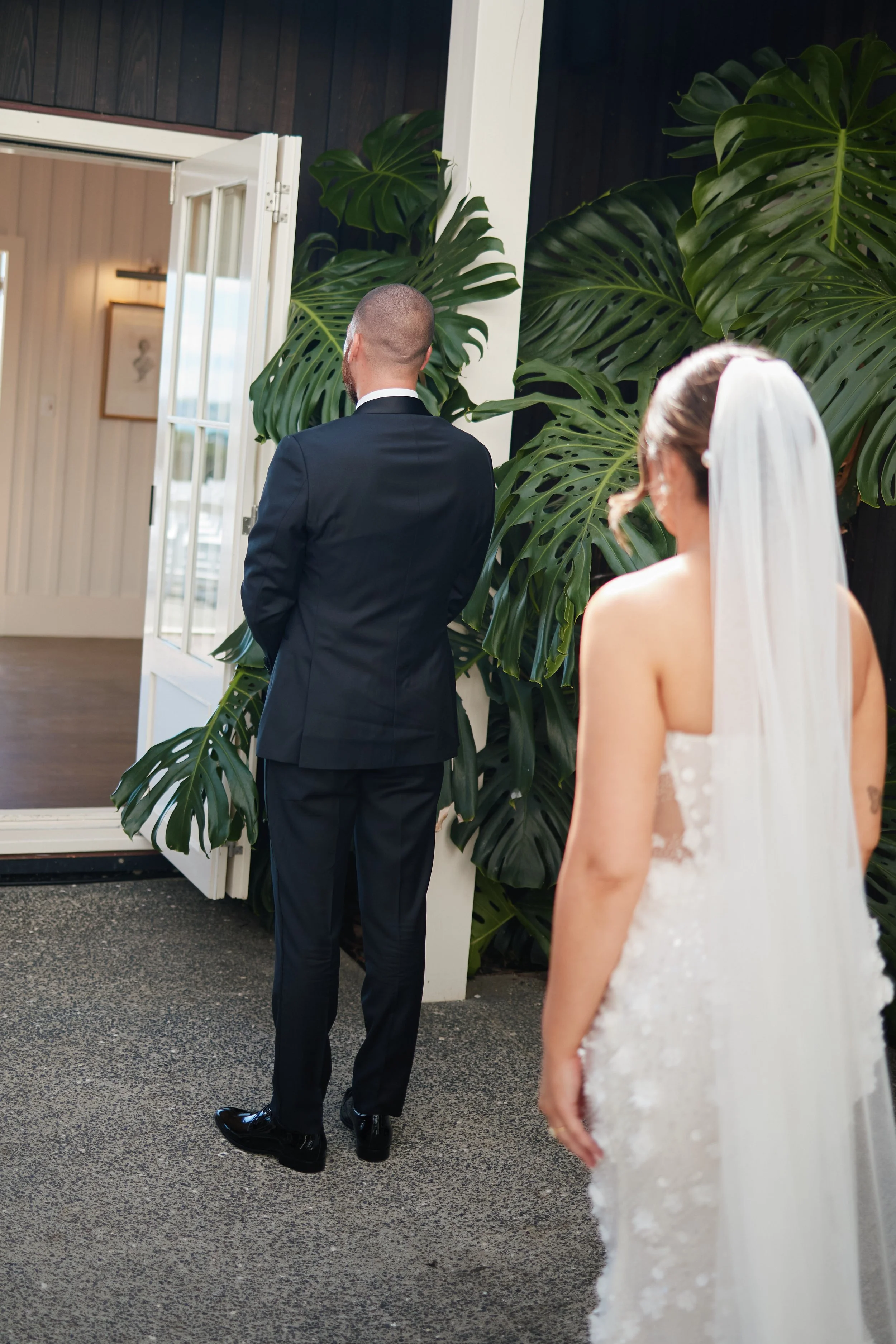 Bride in a white wedding dress with a veil watching groom in a black suit looking out a door, with large green plants nearby.