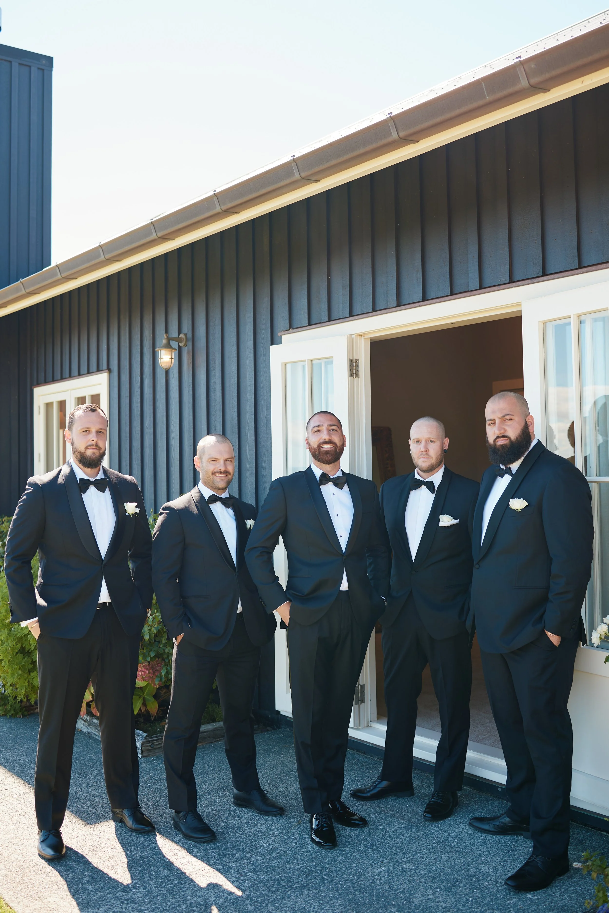 Group of five men in tuxedos standing outside near a house, smiling.