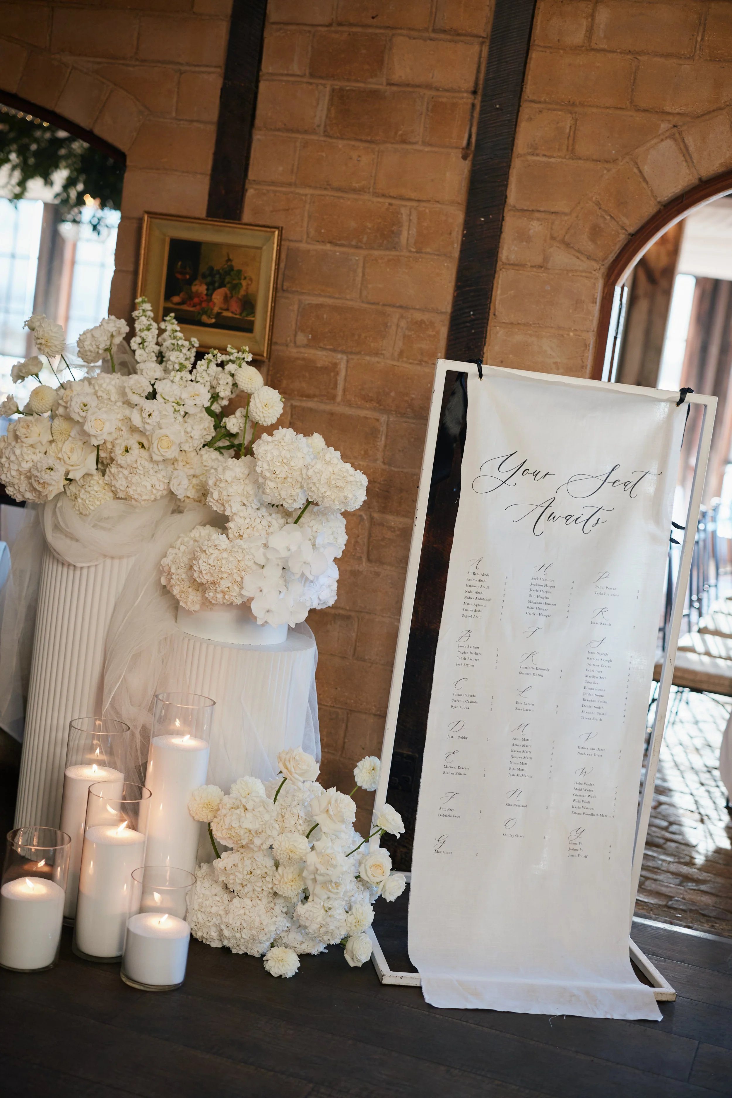 Wedding seating chart sign beside a flower arrangement with white flowers and candles in glass holders at a wedding reception.