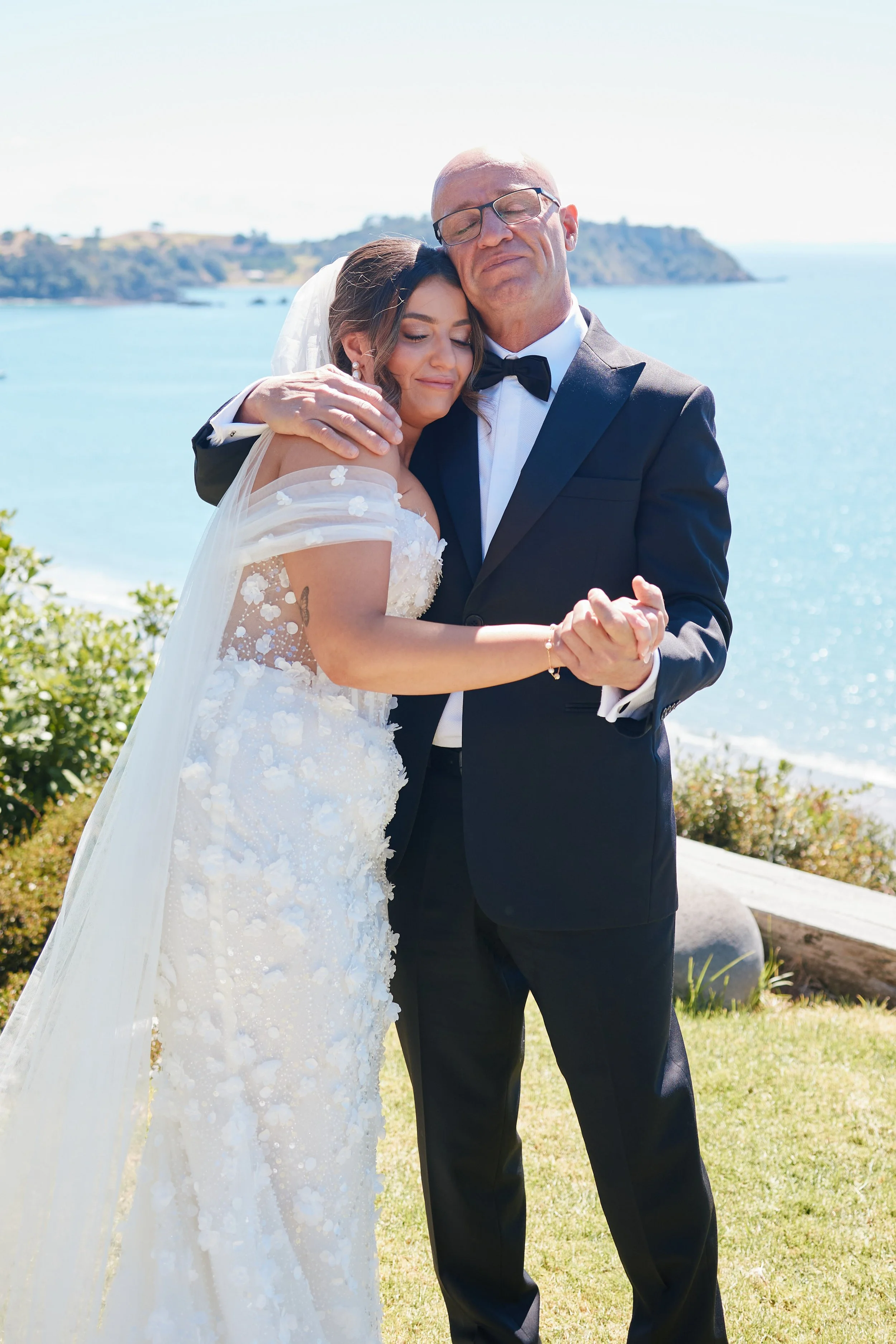 A bride and an older man, possibly her father, dancing outdoors near the coast during daytime.