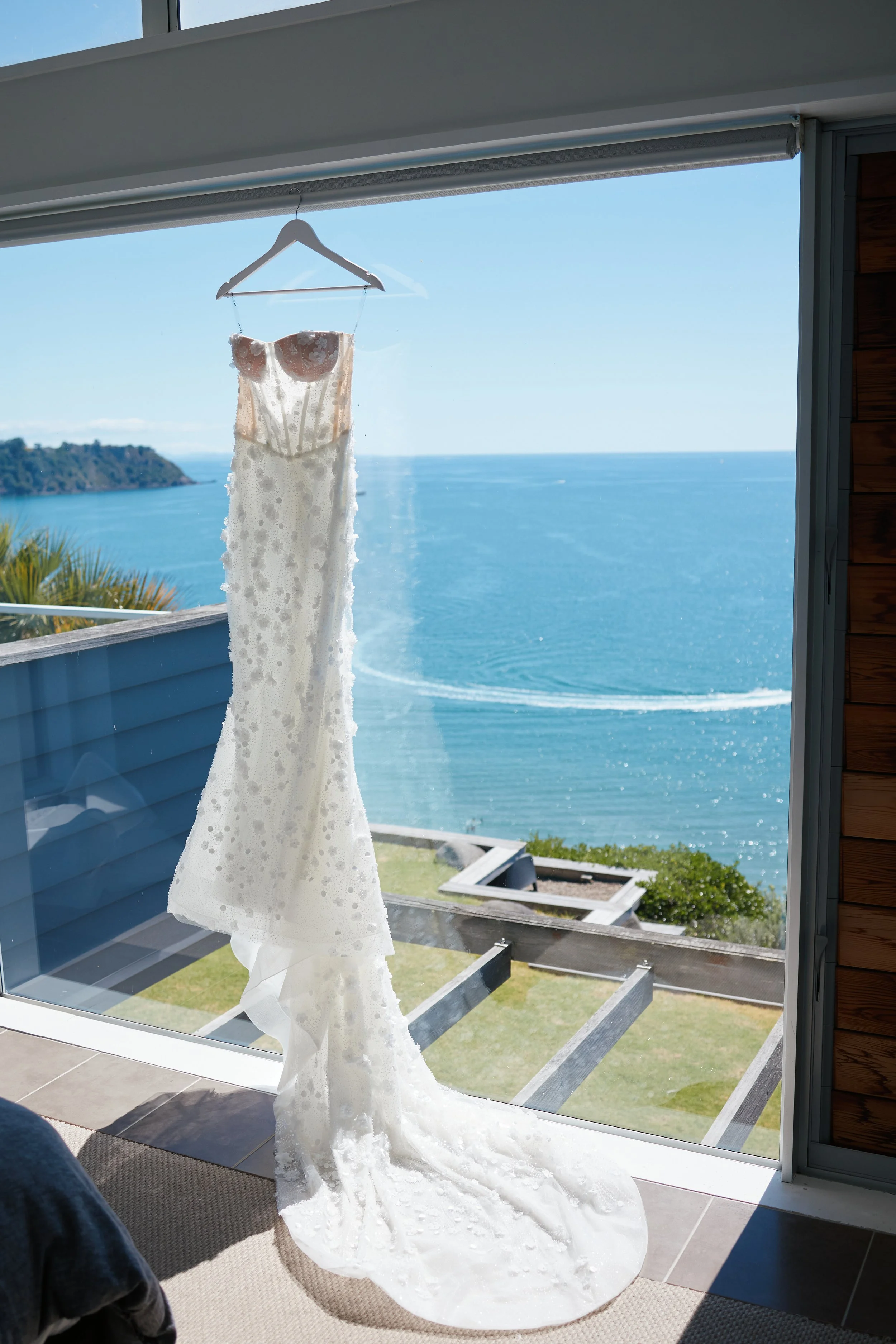 A white wedding dress hangs on a hanger inside a room with a panoramic ocean view in the background.